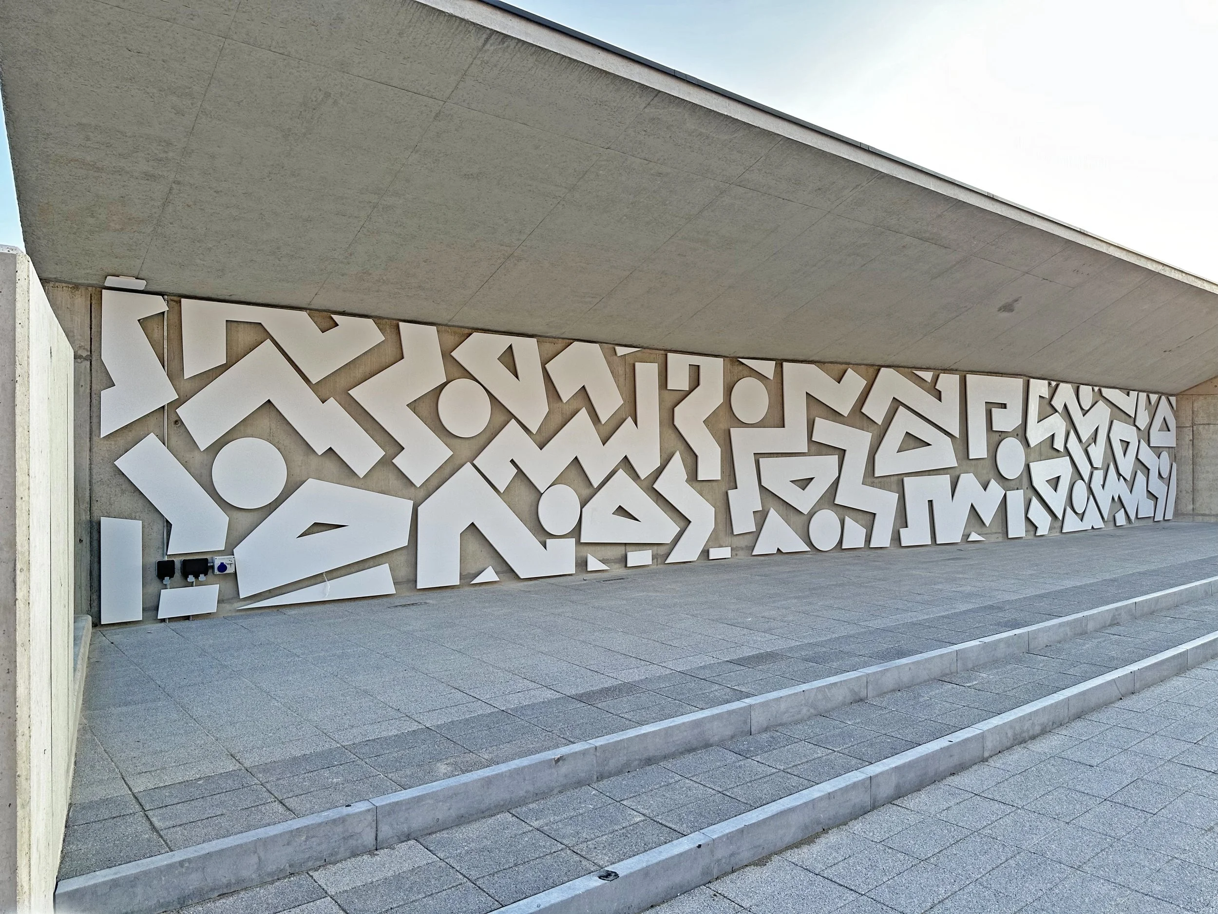 Exterior wall with modern white geometric abstract art installation beneath a concrete ceiling, surrounded by a paved area with steps