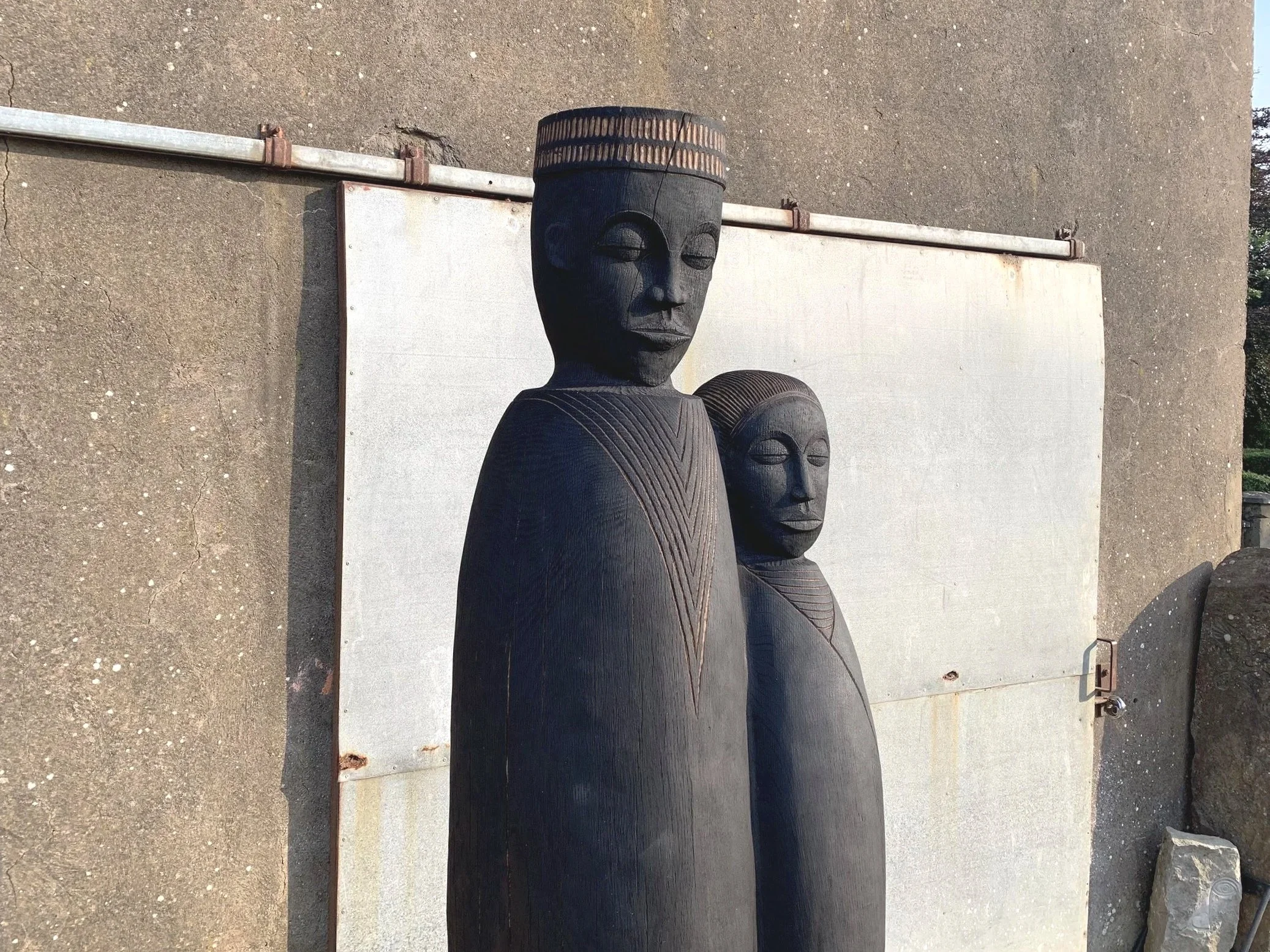 Two tall black wooden sculptures of human figures with elongated faces, set outdoors against a concrete wall with a white panel, surrounded by rocks and urban elements.