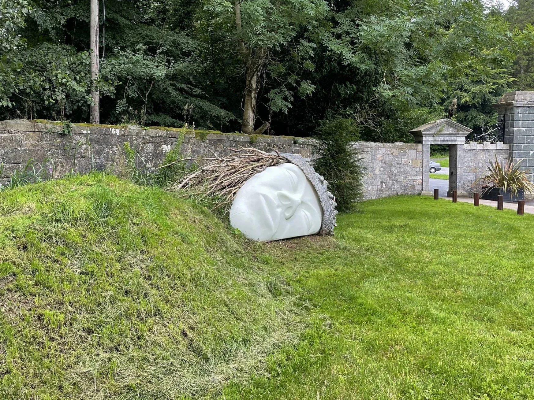 A large sculpture of a person's face with closed eyes, lying on a grassy slope and covered with branches and leaves, against a stone wall and lush trees.
