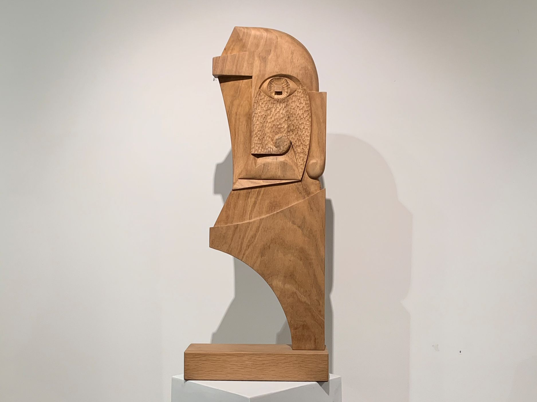 Abstract wooden sculpture of a stylized human face with geometric features, displayed on a white pedestal against a light-colored wall.