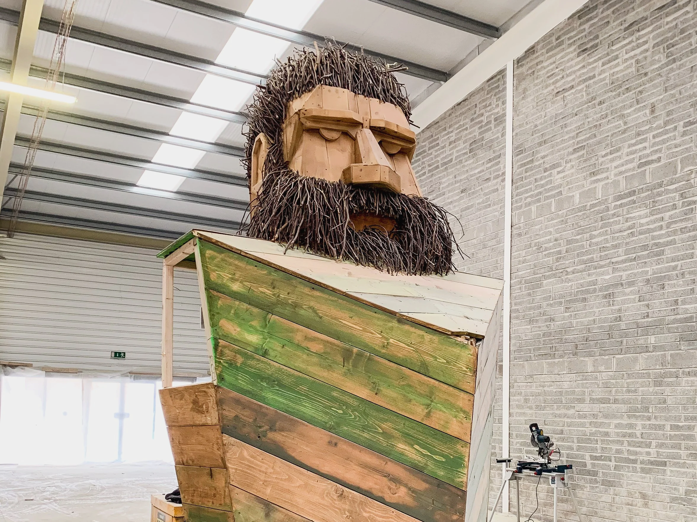 A large sculpture of a man with a beard, made of wood and branches, showing a serious expression, inside a warehouse or workshop.
