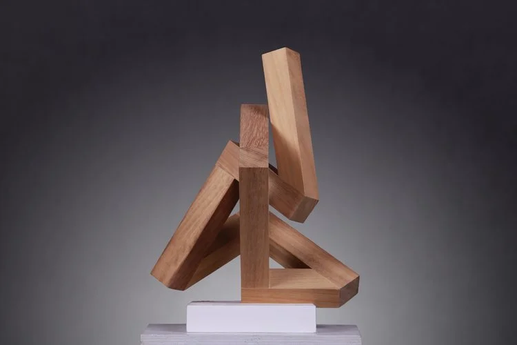 Wooden block sculpture assembled in an abstract, geometric design, placed on a white base against a dark background.