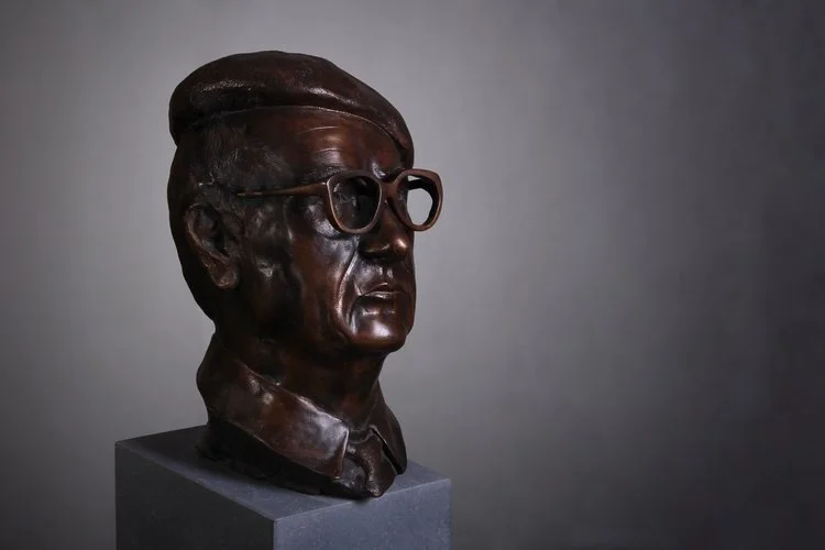 Bronze bust of a man wearing glasses and a beret, displayed on a pedestal against a gray background.