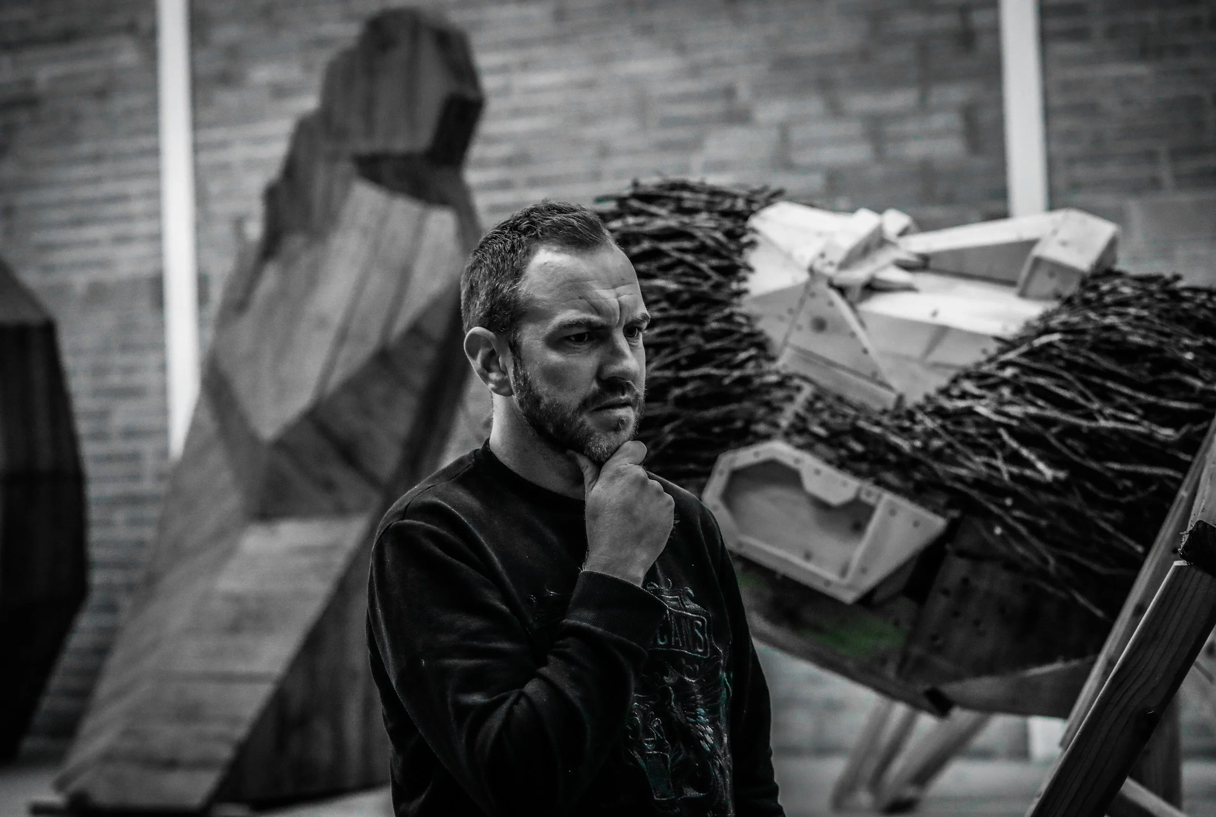 A man with a beard and short hair, wearing a dark sweatshirt, is standing in front of wooden and mixed media sculptures in an art studio or gallery, with his hand on his chin in a thinking pose.