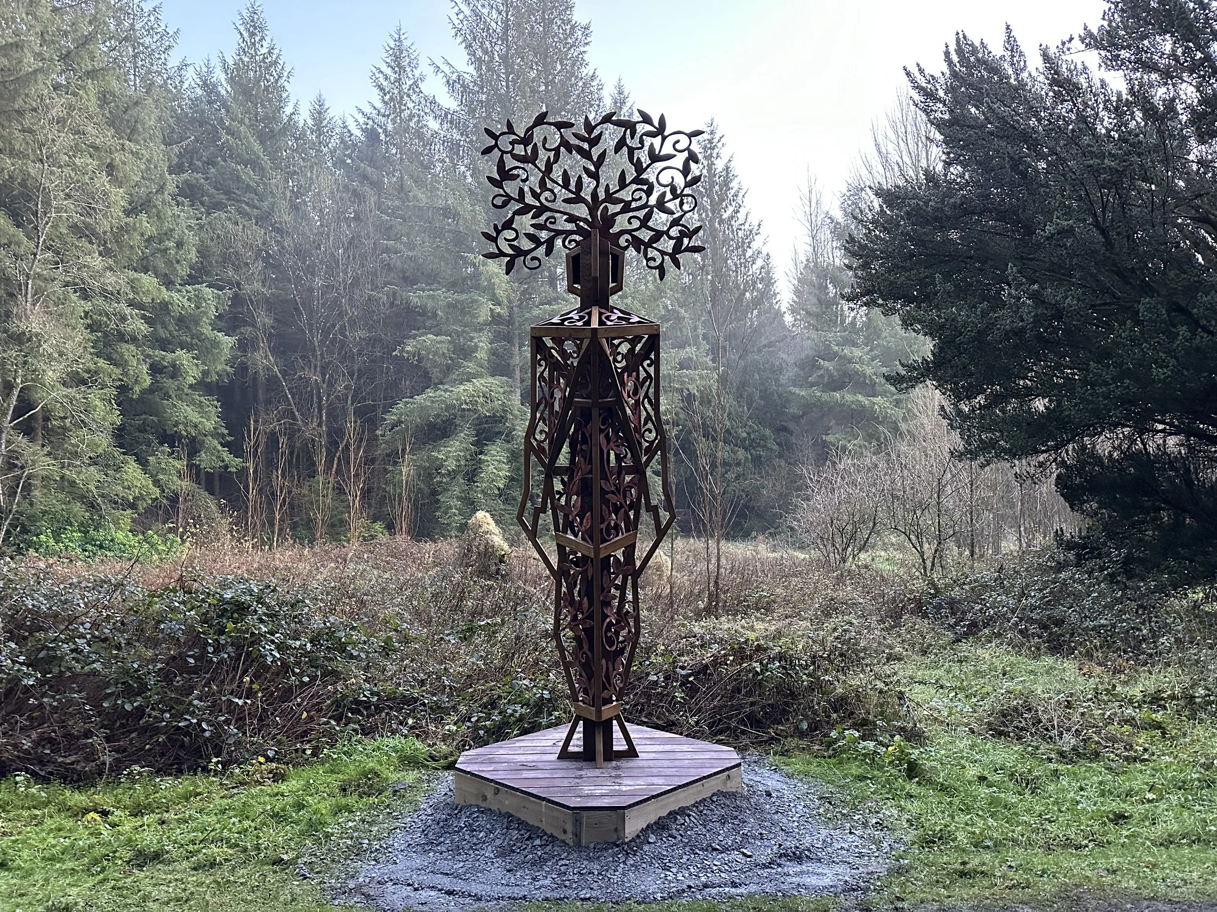 A decorative metal sculpture of a tree mounted on a wooden platform in a forest clearing.