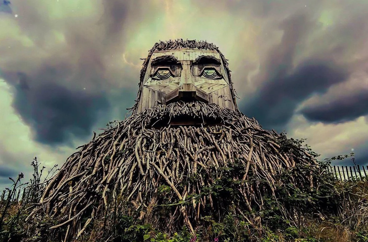 A giant sculpture of a bearded man with a face made from wood and metal, set against a cloudy sky.