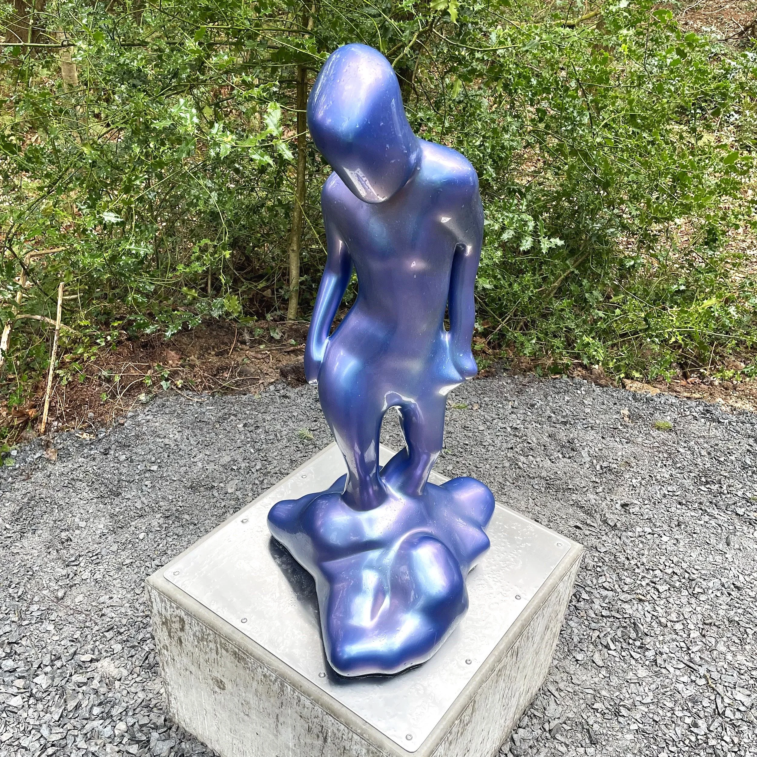 Sculpture of an abstract, glossy, blue figure with a distorted, elongated form standing on a square concrete base outdoors, surrounded by green bushes and gravel ground.