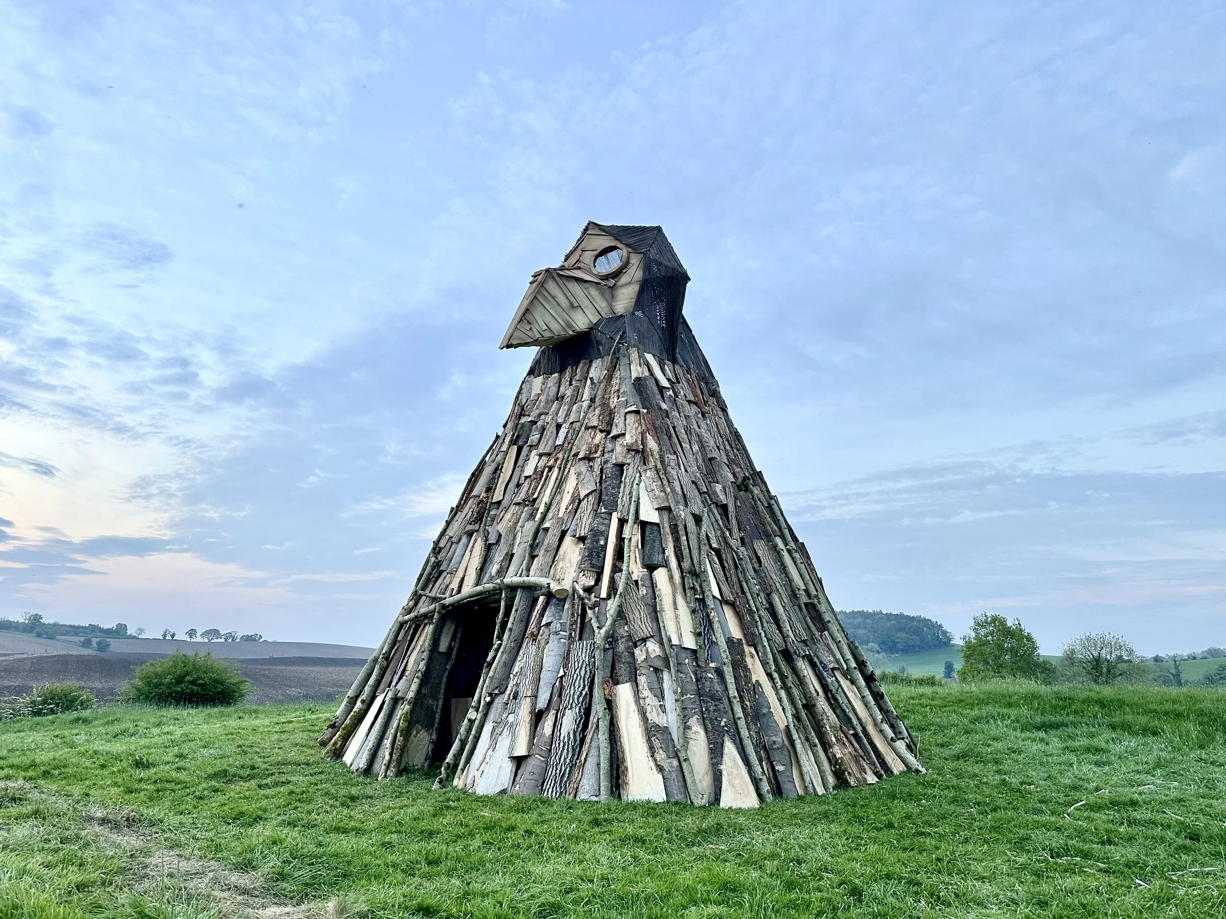 A large, cone-shaped wooden structure resembling a bird or a creature with a beak, made of stacked wooden planks, situated in an open grassy field with hills and trees in the background under a partly cloudy sky.