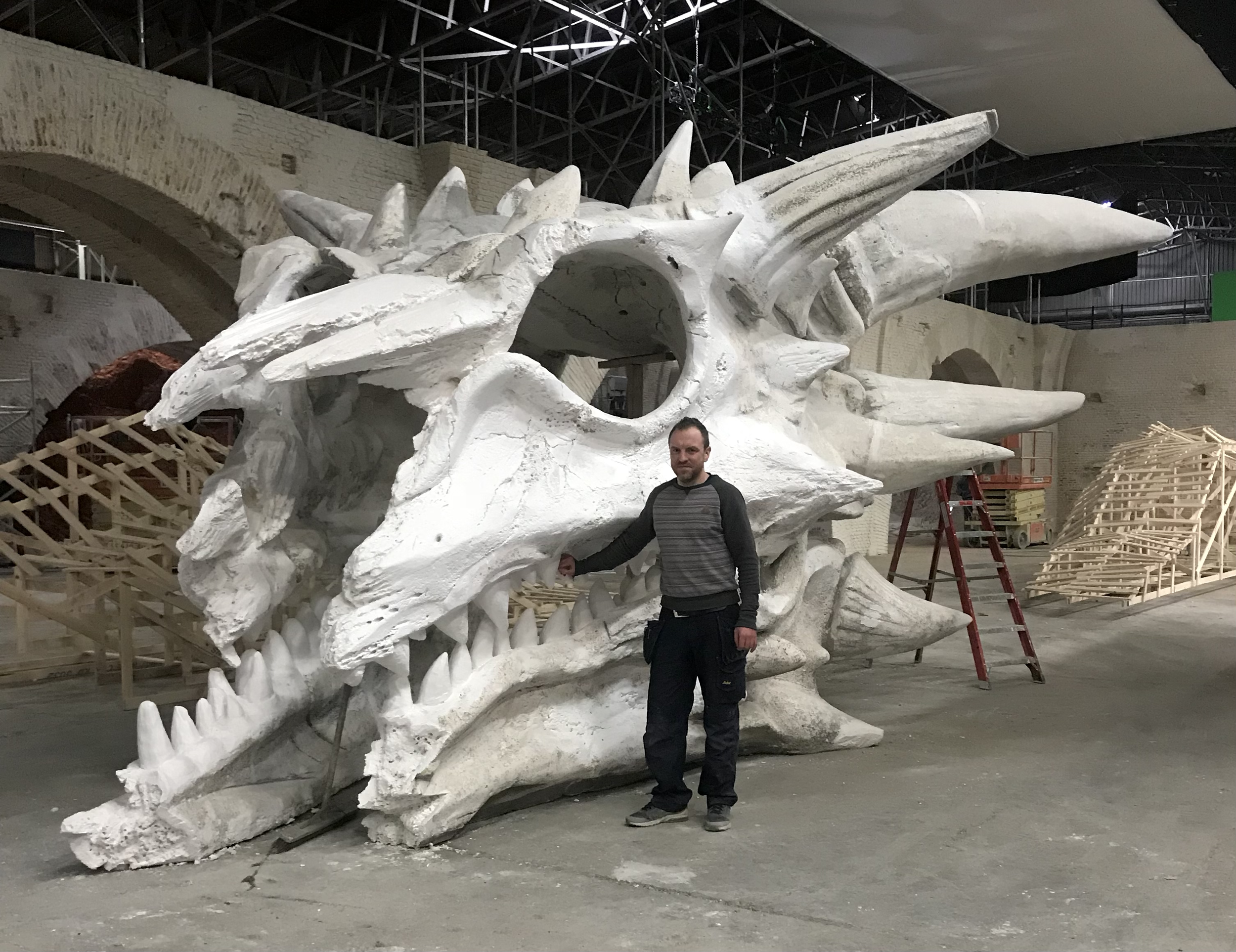 A man standing next to a large, white dragon skull sculpture under construction in a workshop.