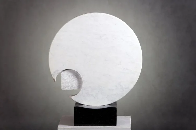 A modern sculpture featuring a large white circle with a smaller cut-out circle on the lower left, mounted on a black rectangular base against a gray background.