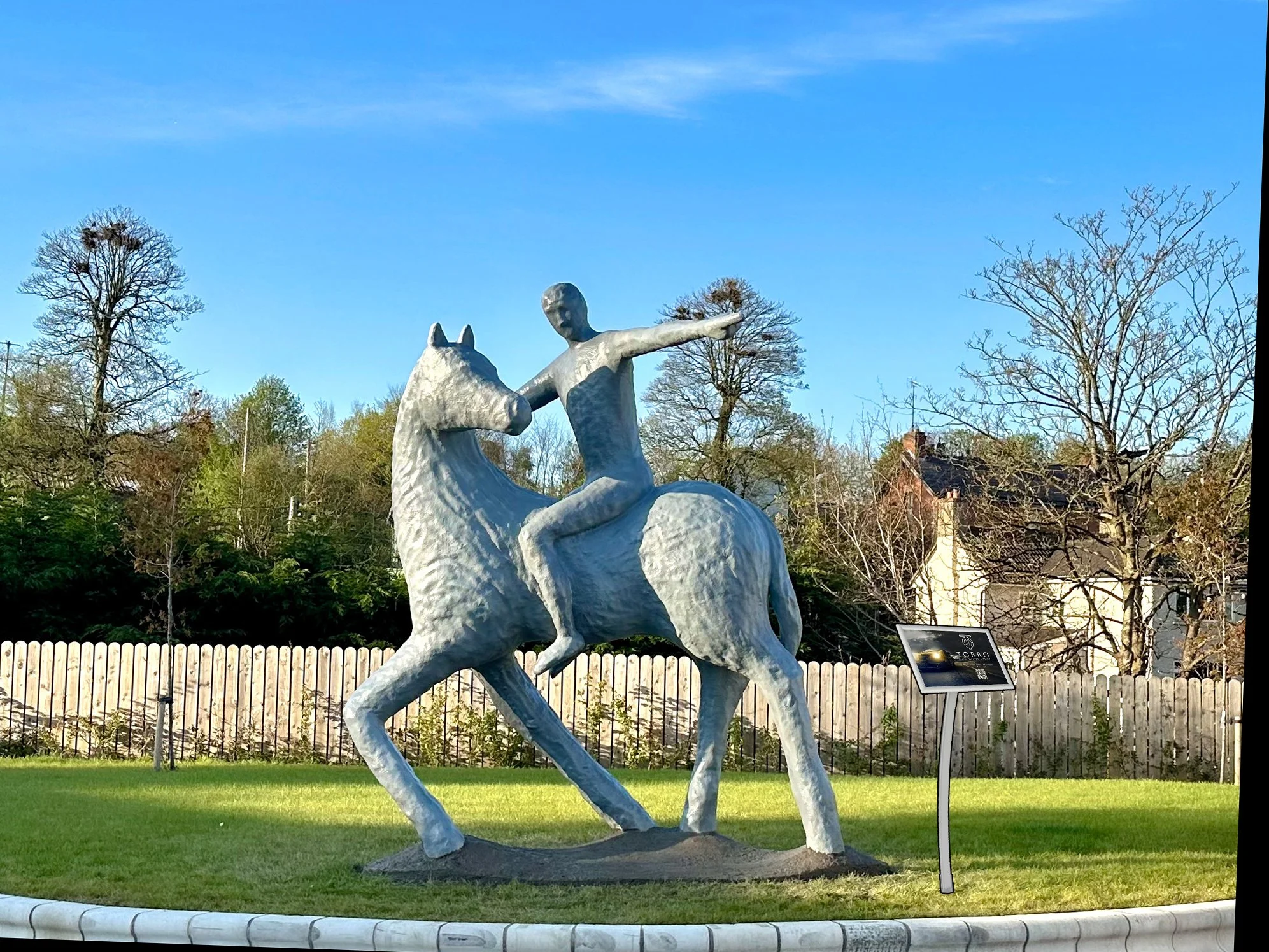 A large outdoor sculpture of a person riding a horse in a park, with trees and a house in the background and a clear blue sky.