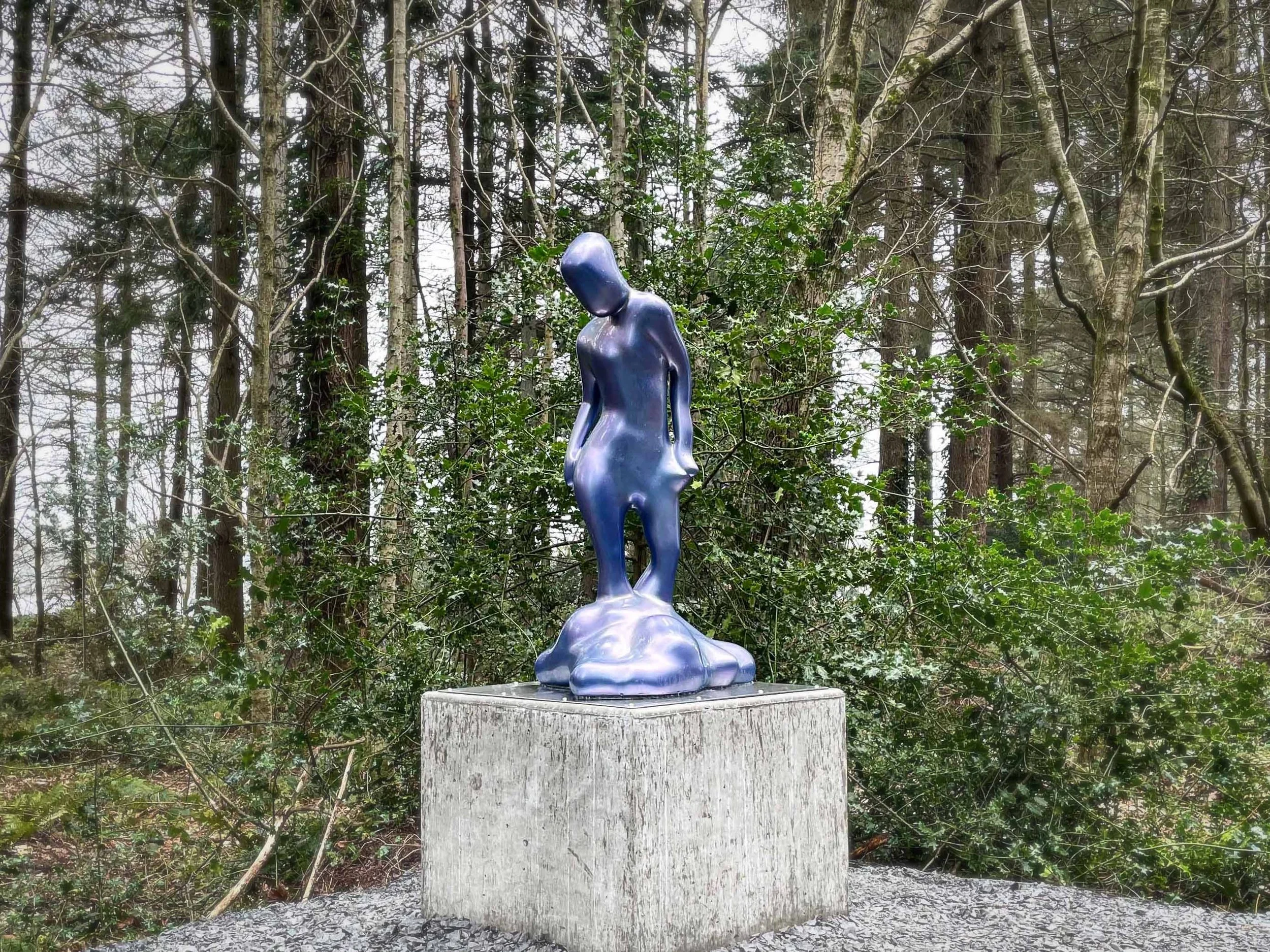 Blue abstract human sculpture on wooden pedestal in a forested area.