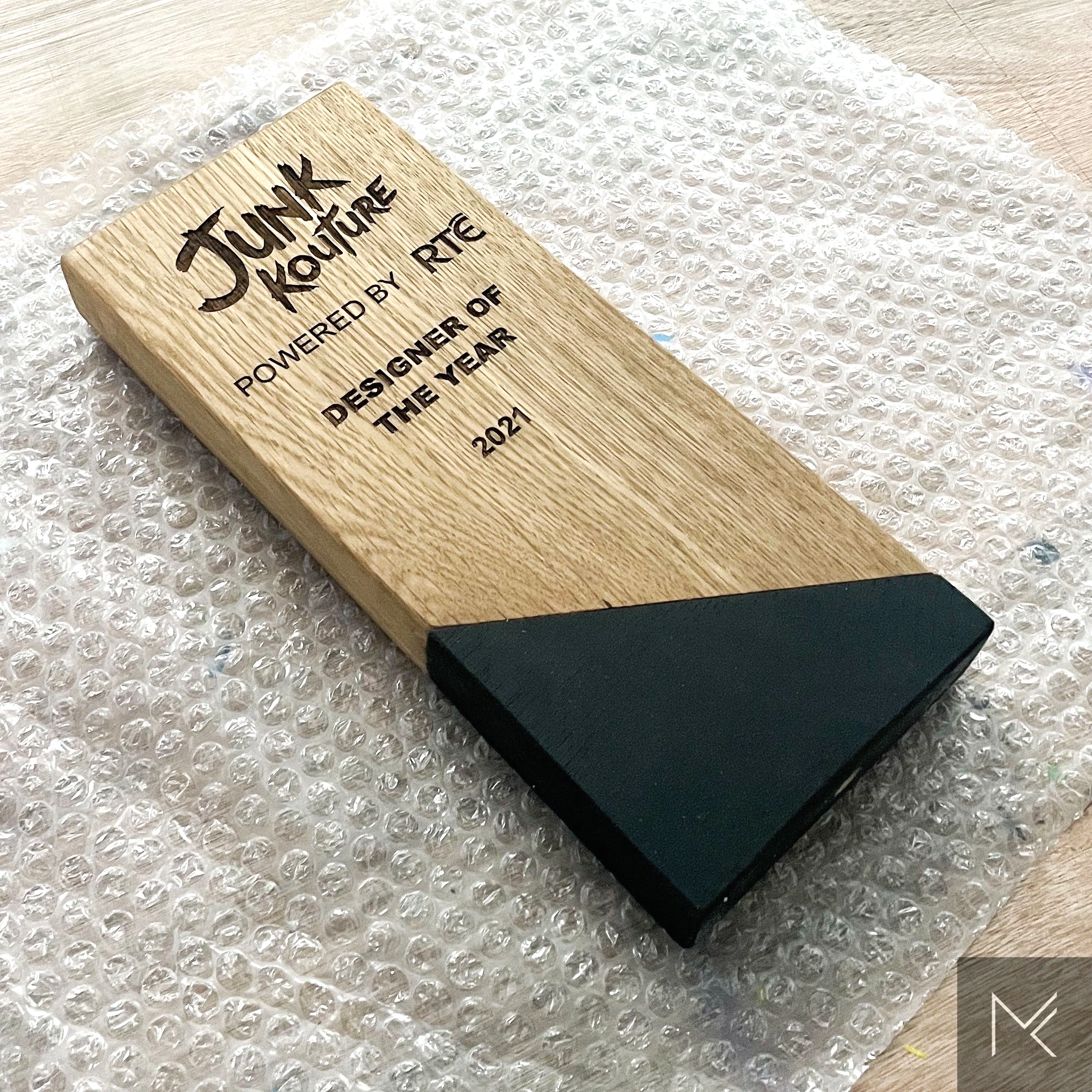 A wooden award plaque with black text reads 'Junk Couture powered by PTEC, designer of the year 2021,' resting on bubble wrap.
