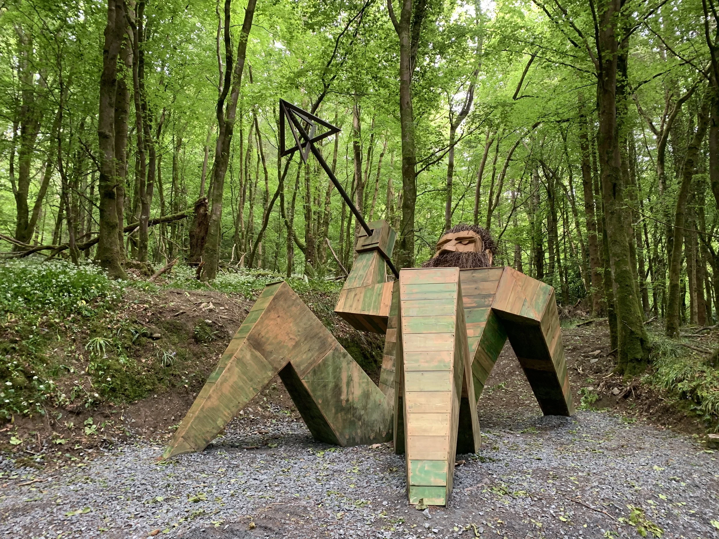 Wooden sculpture in the shape of a creature with a rock-like face and a triangular antenna, situated in a lush green forest