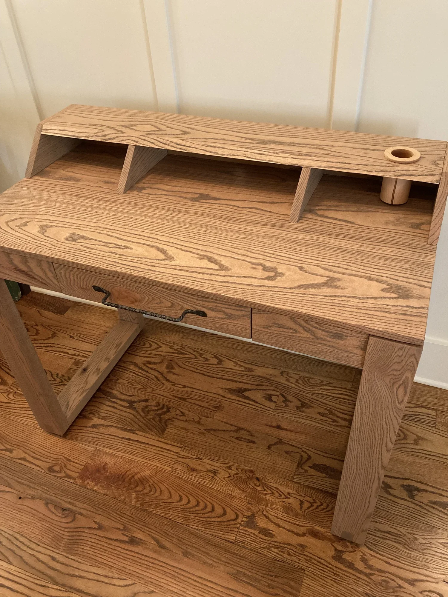 A wooden desk with a top shelf and a small hole on the right side, likely for holding a pen or pencil, sitting against a white wall with wooden flooring.