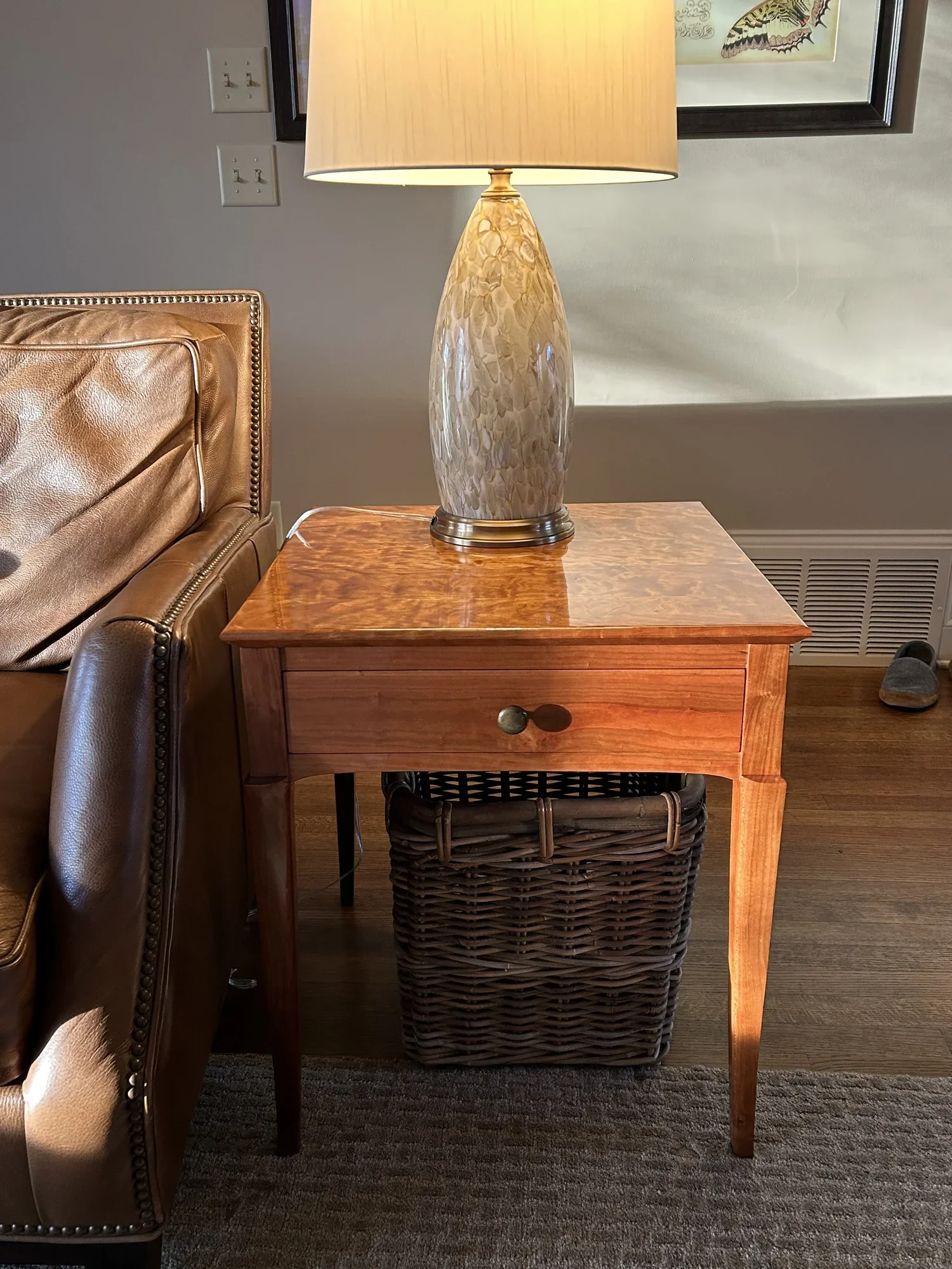 A wooden side table with a drawer, beneath which hangs a woven basket. On top of the table is a large ceramic lamp with beige shade. Part of a leather couch is visible to the left. The background has a framed picture and a wall outlet.