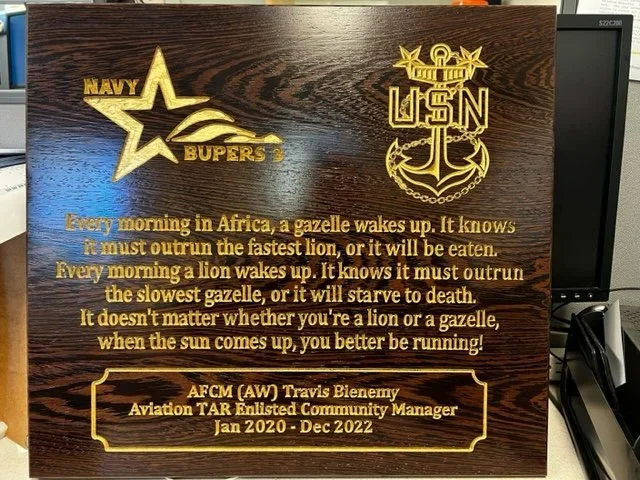 A wooden plaque engraved with a Navy star logo and the words "Navy Buds" on top, along with a USN emblem. Below is a quote about lions and gazelles, and at the bottom, it commemorates AFCM (AW) Travis Bienemy, an aviation TAR Enlisted Community Manag