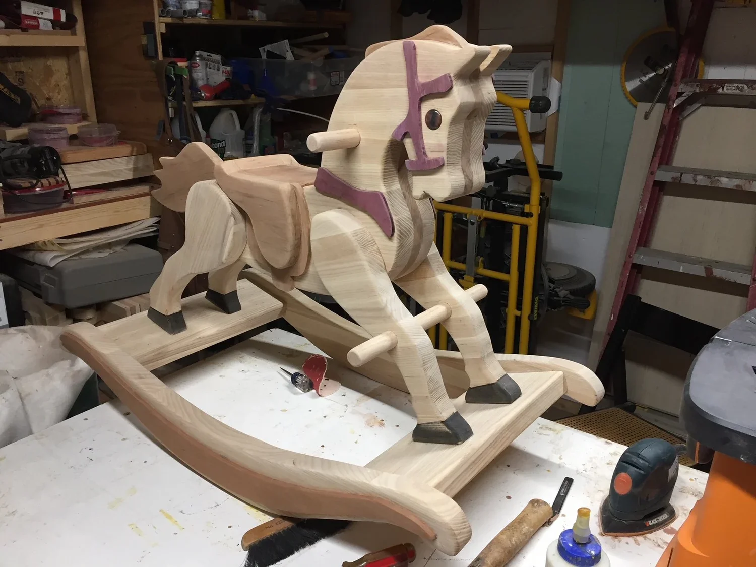 A handcrafted wooden rocking horse in a workshop, unfinished, with visible wood grain and parts assembled, surrounded by tools and materials.