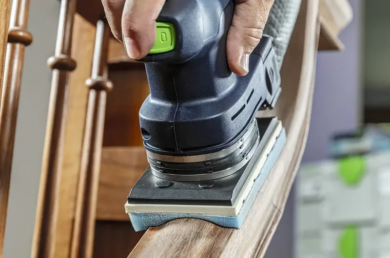 Close-up of a person using a Festool sander on a wooden surface, with a background of furniture and household items.
