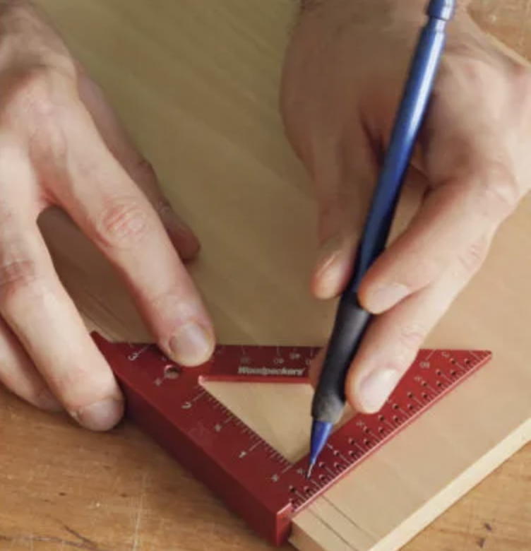 A person is using a blue pen and a Woodpeckers square to draw a straight line on a wooden surface.