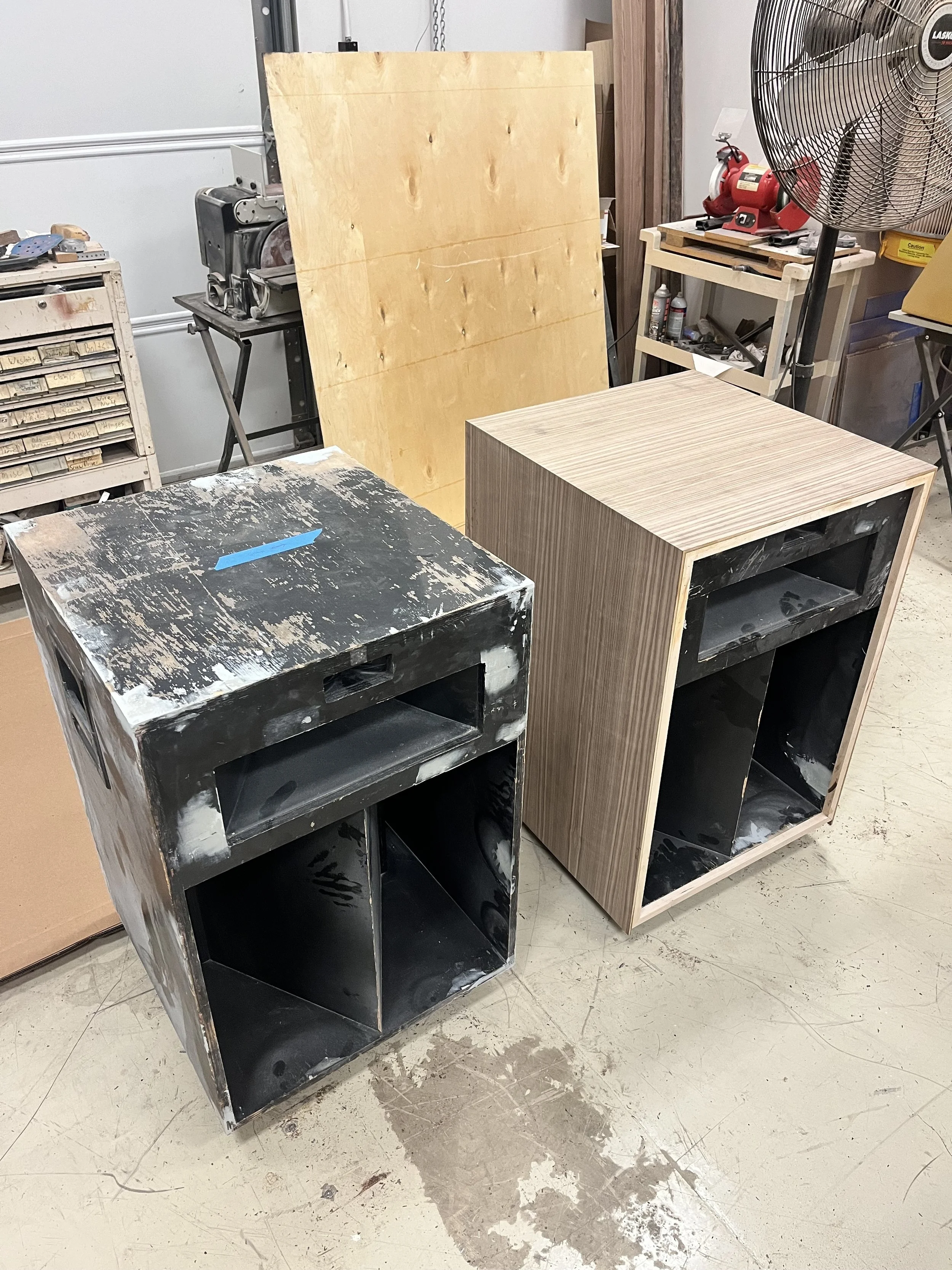 A before and after of unfinished wooden speaker cabinets in The Woodwork Shop, one with a black painted interior, the other with a light wood veneer exterior, surrounded by woodworking tools and equipment.