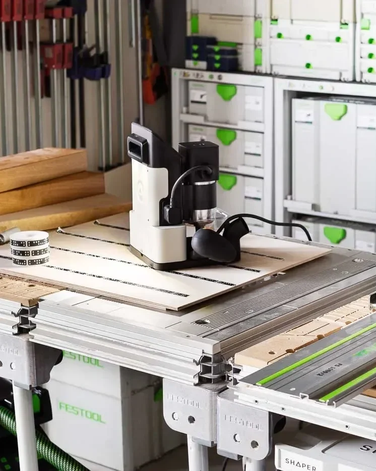 A woodworking workshop with a Shaper Origin on a worktable, surrounded by wood pieces, clamps, and systainers.