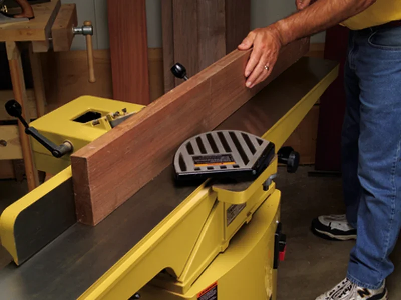 A person using a Powermatic jointer to flatten a wooden board in a woodworking shop.