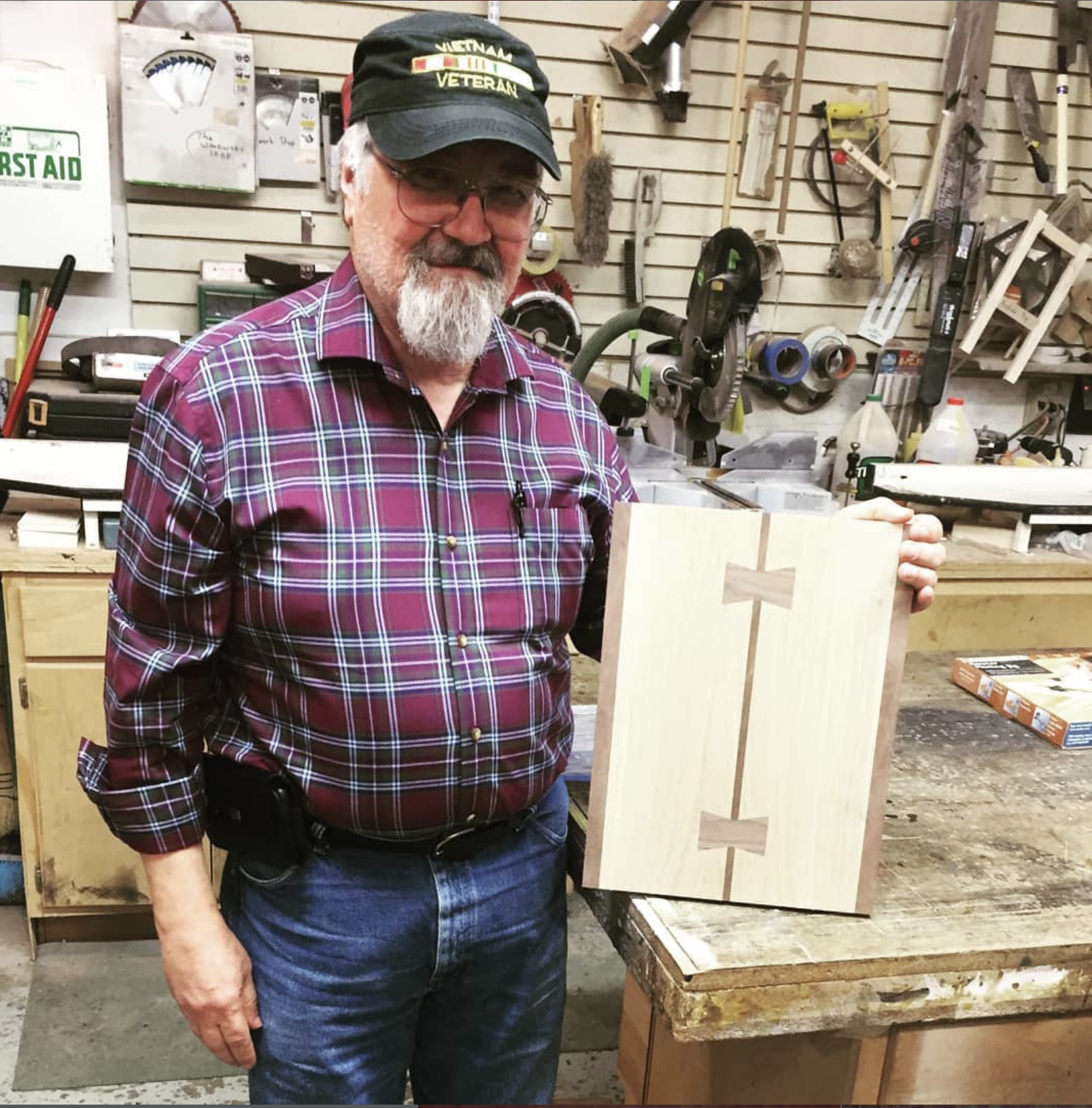 A man with glasses, a beard, and a Veterans cap holding a piece of wood with a religious cross design at The Woodwork Shop. 