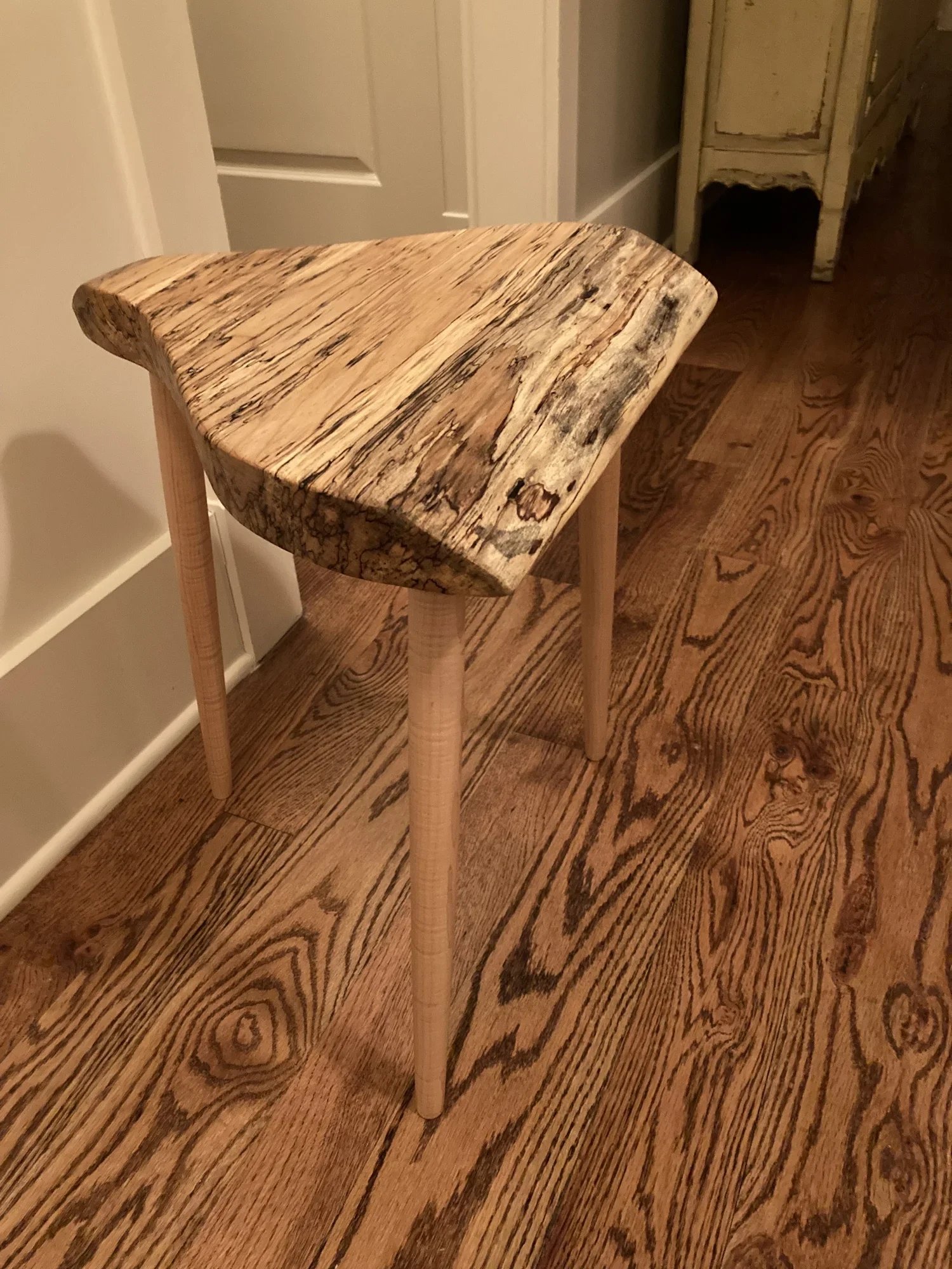 A small wooden table with a natural, uneven, and rough-cut wood top and three light-colored wooden legs, situated on a wooden floor near a wall and a door.