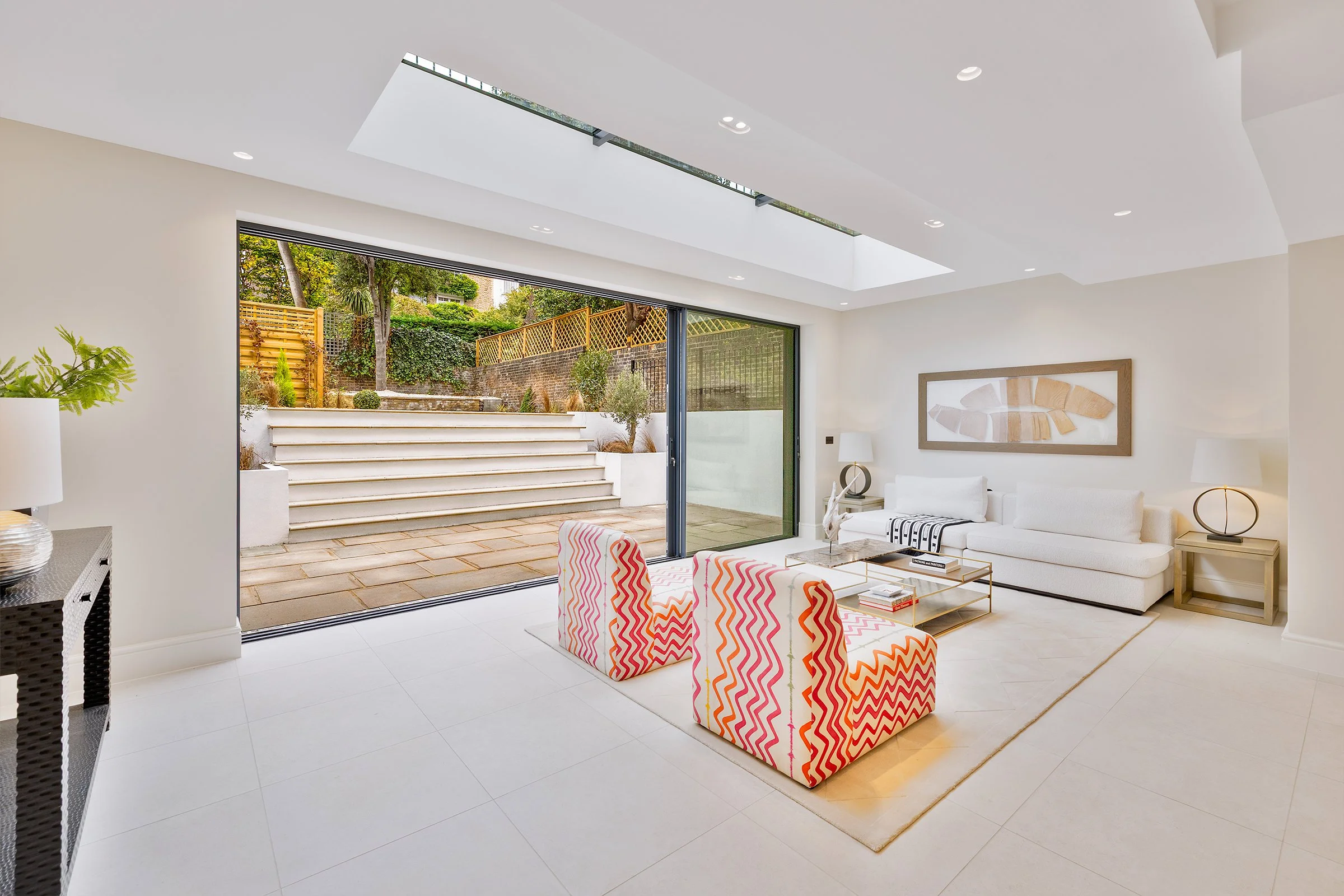 Bright living room with white walls and tiled floor, large glass sliding door opening to patio with steps and greenery, white sofa, colorful patterned chairs, modern lighting and artwork.