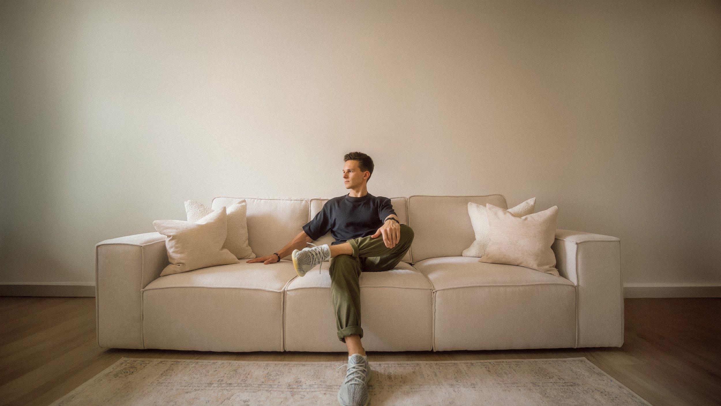 Young man sitting on a white sofa with beige pillows in a minimalist room with a blank wall.