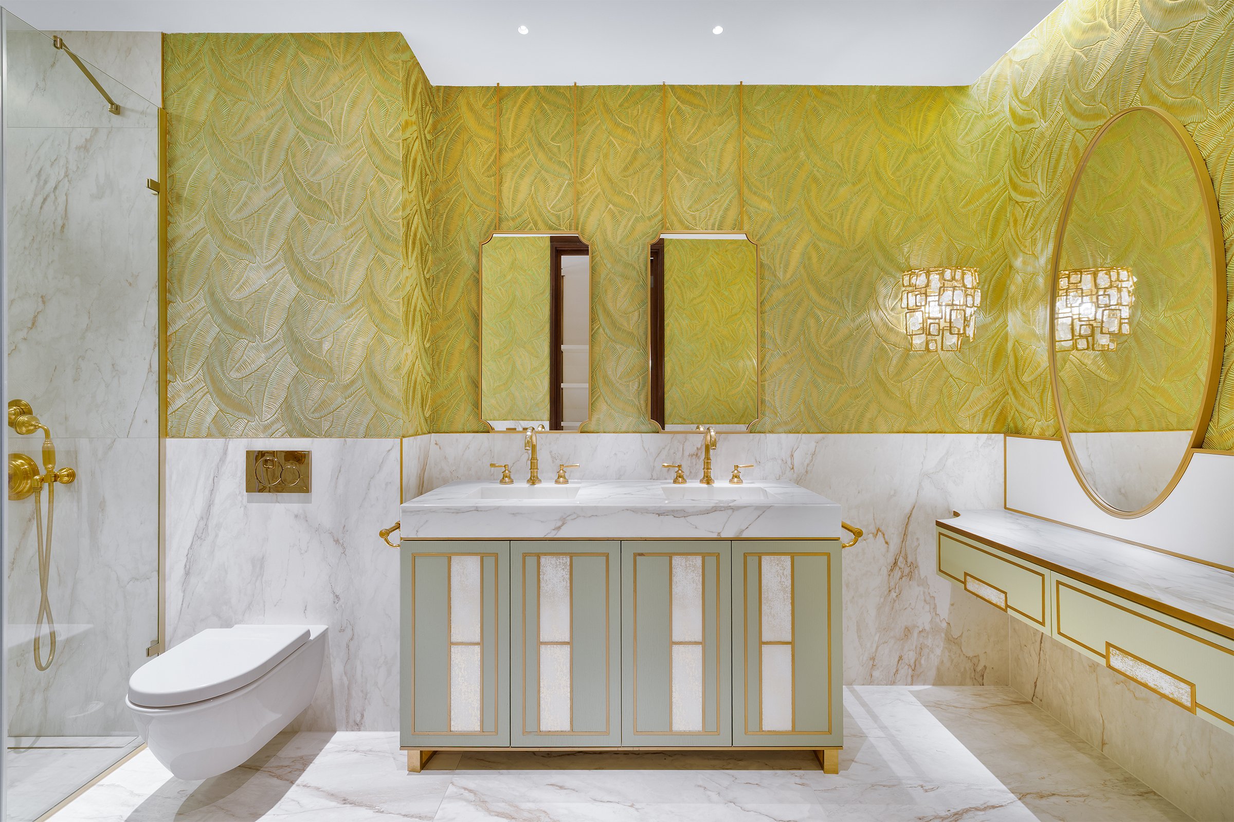 Luxurious bathroom with gold accents, green patterned wallpaper, white marble sink vanity, large mirror, and a round mirror, with a toilet and a glass-enclosed shower area.