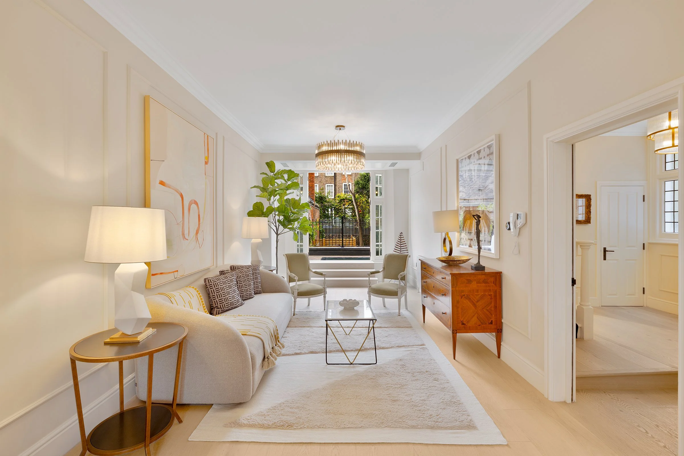 Bright living room with cream-colored sofa, two armchairs, a small coffee table, a large potted plant, wooden sideboard, artwork on walls, and an open window showing outdoor greenery.