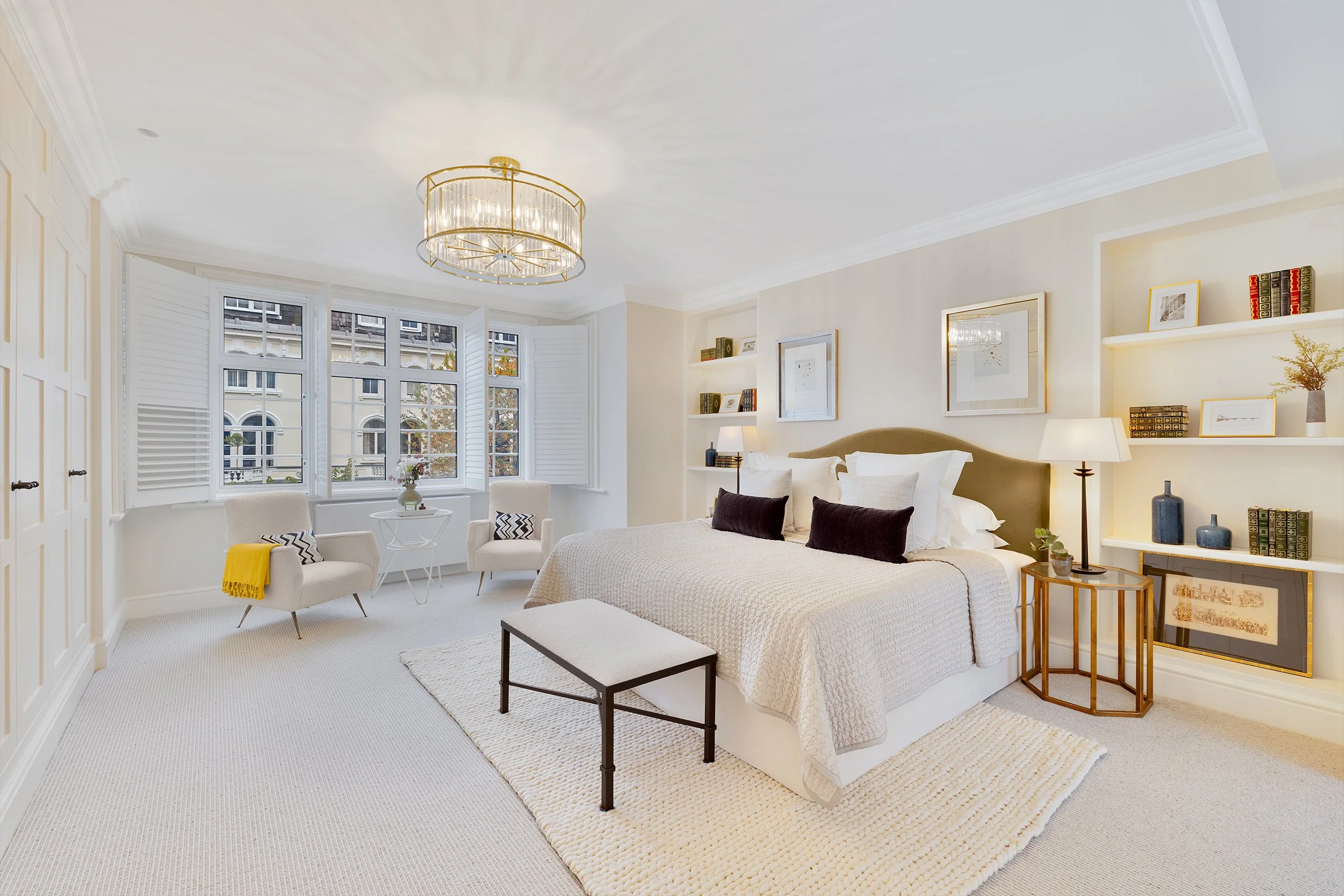 Bright bedroom with large window, beige walls, white ceiling, round chandelier, bed with white and black pillows, white bedding, beige headboard, white built-in bookshelves with decorative items, white armchairs, small round table with flower, bedsid