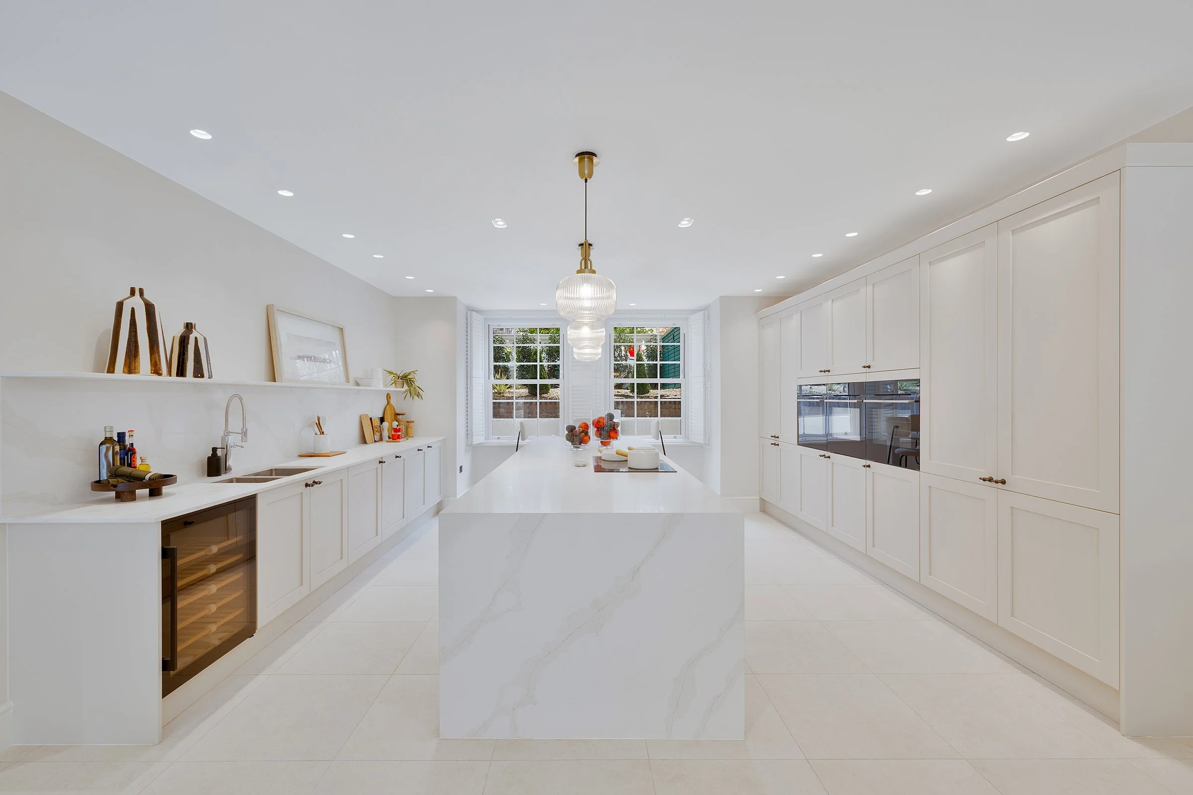 Bright, modern kitchen with white cabinets, a large white marble island, and a row of recessed ceiling lights. There is a chandelier over the island and a window at the end showing outdoor greenery.