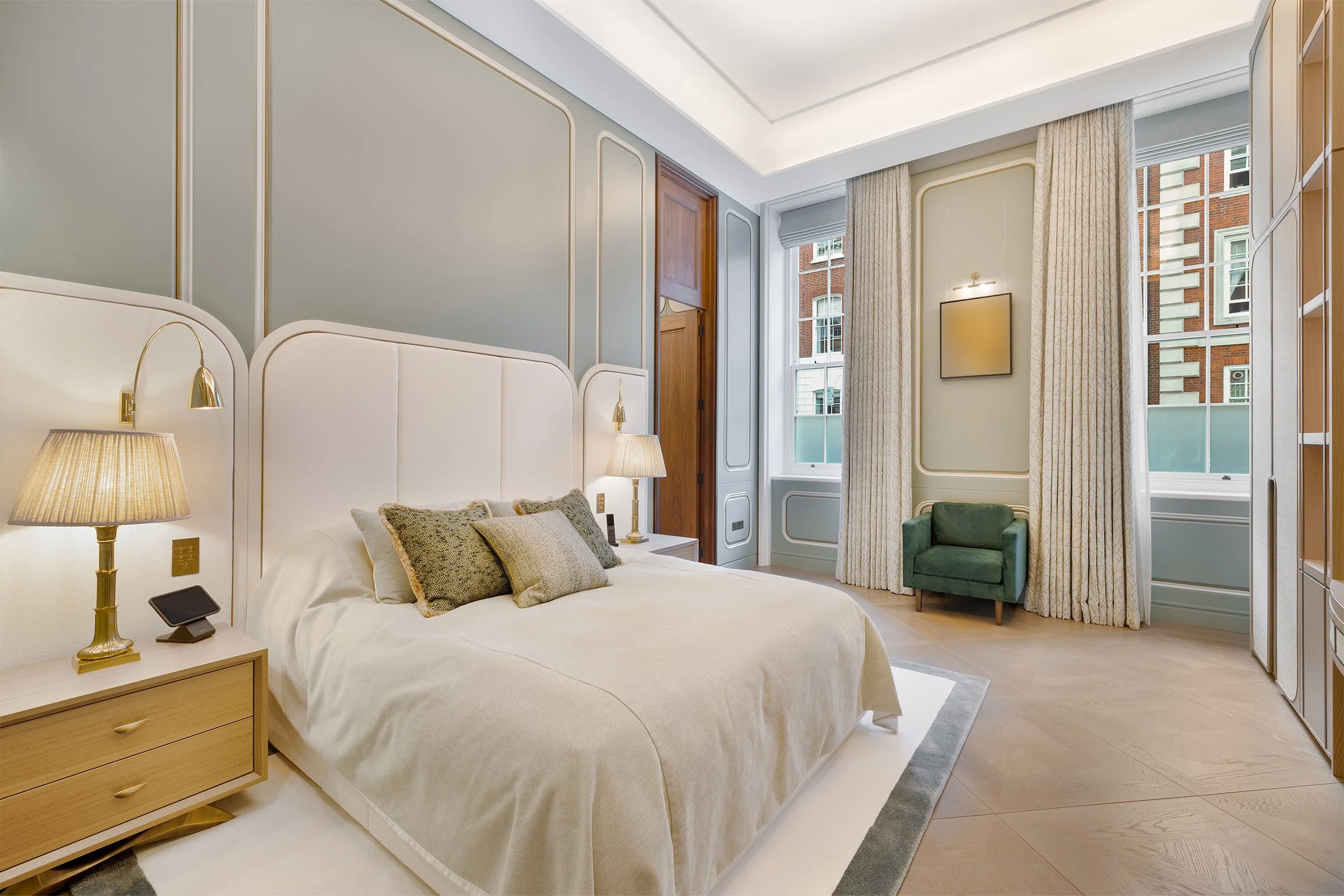 A modern bedroom with a large bed, green accent chair, wooden nightstand with lamp, and large windows with curtains, overlooking a brick building.