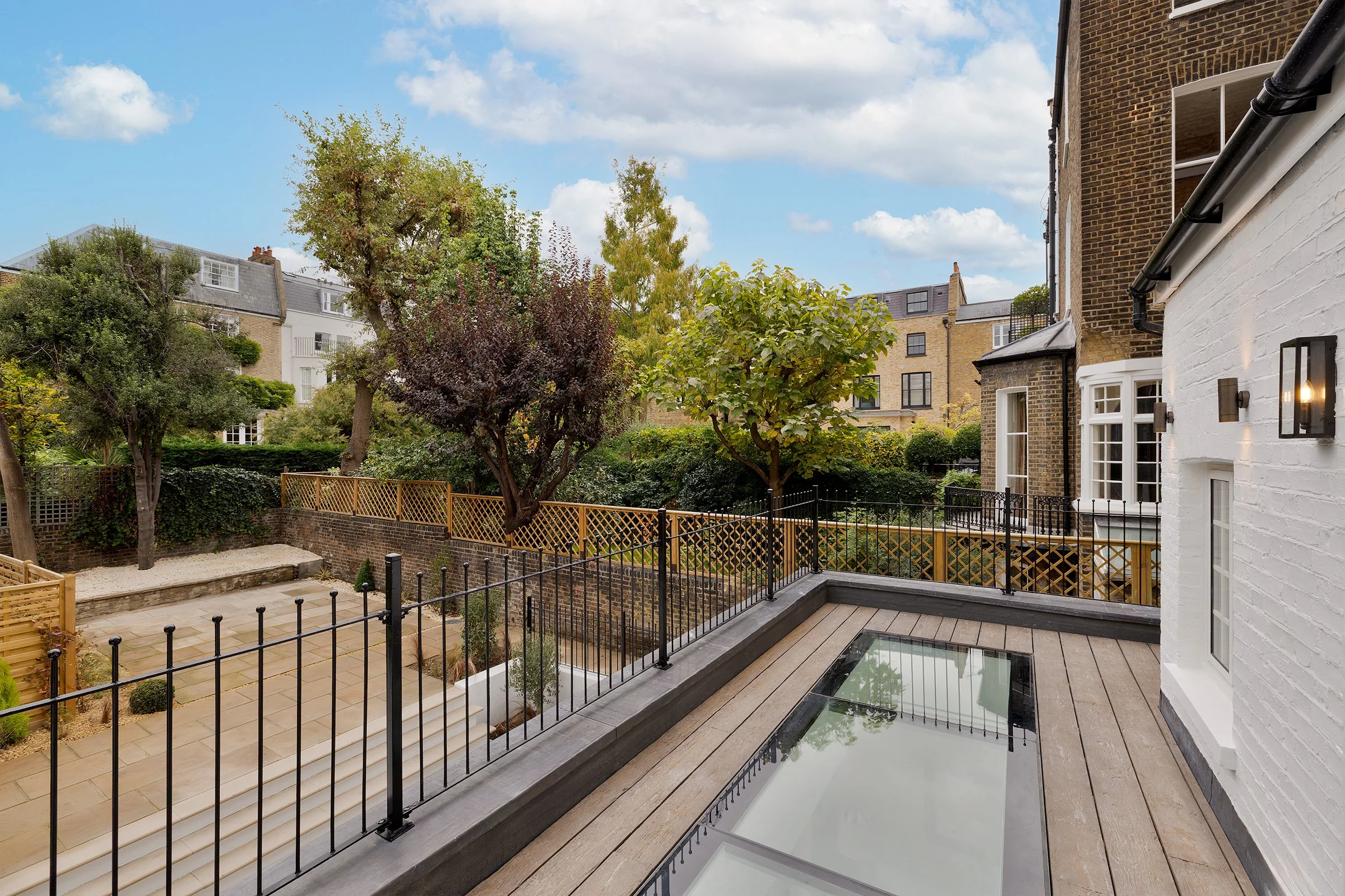 View of a backyard patio with wooden decking, glass skylight, black metal fence, trees, bushes, and neighboring houses under a partly cloudy sky.