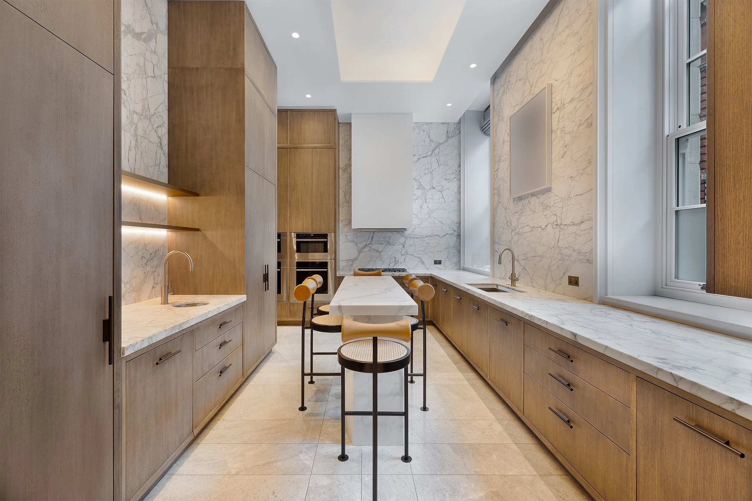 Modern kitchen with wooden cabinetry, marble countertops, and a marble backsplash. There are large windows on the right side and a small kitchen island with stools in the center.