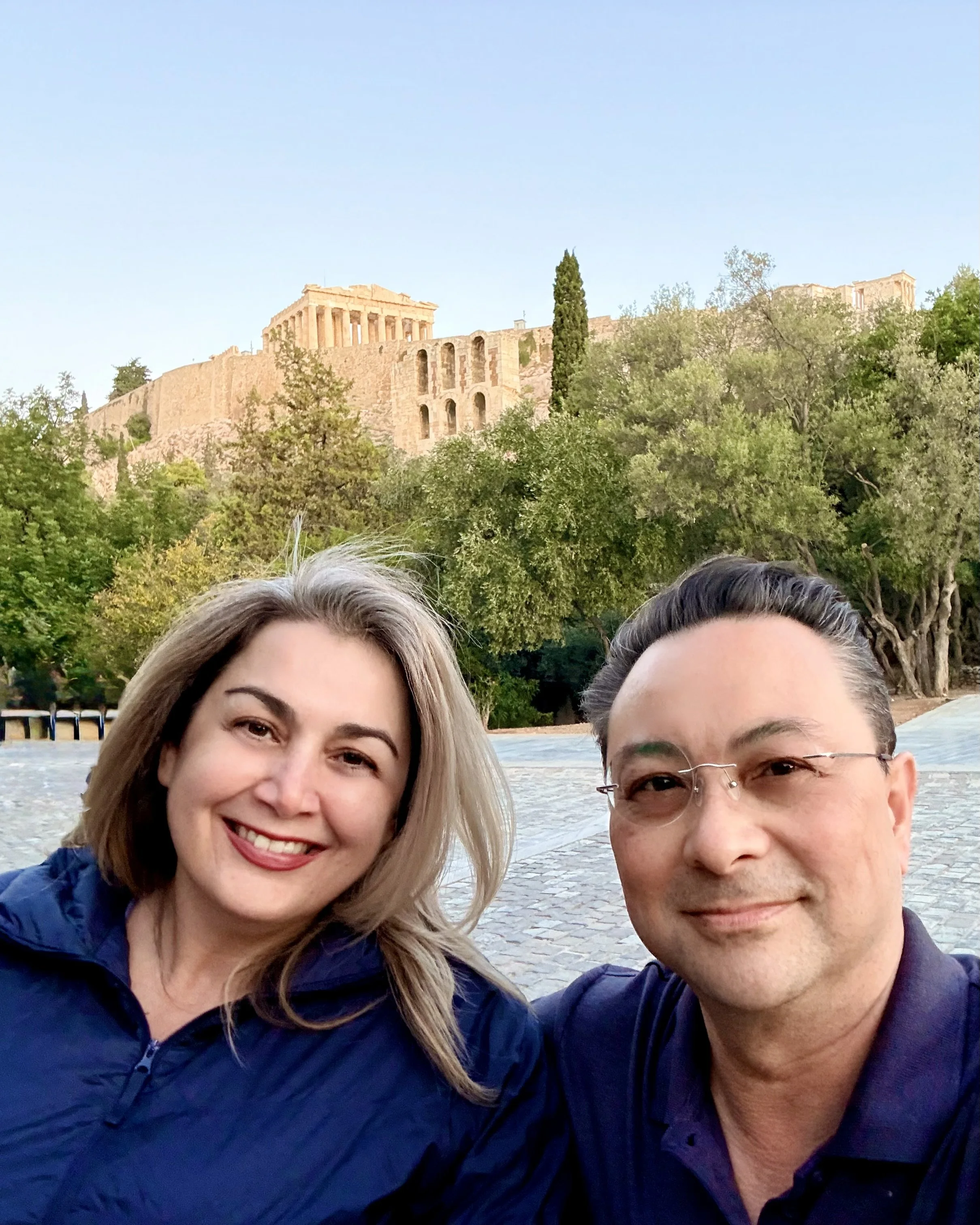 A smiling woman with blonde hair and a man with glasses taking a selfie outdoors. In the background, the Acropolis in Athens, Greece, is visible with its ancient ruins atop a hill surrounded by trees and greenery.