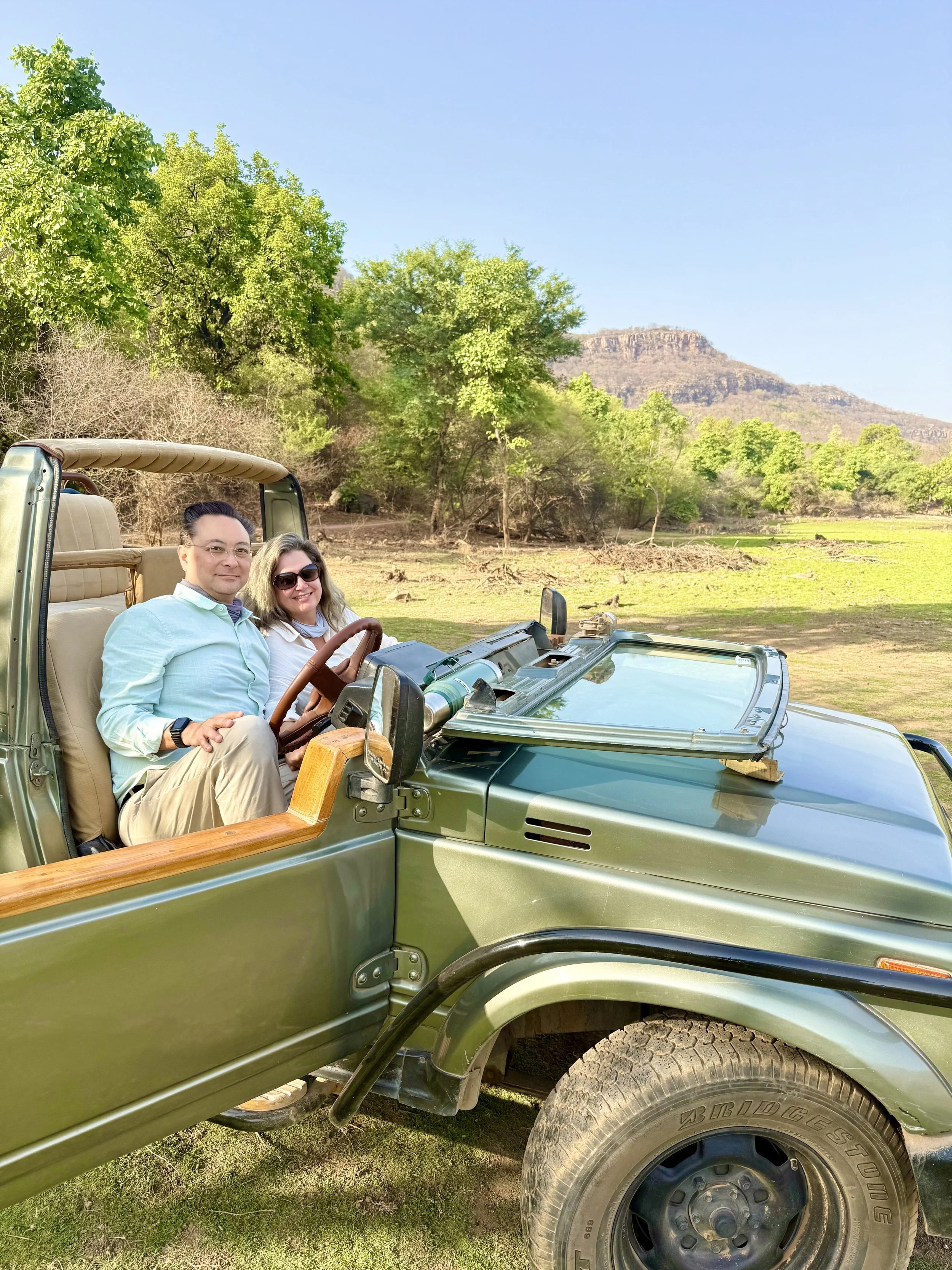 Two people sitting in the front of a green safari vehicle in a scenic outdoor setting with trees and hills in the background.
