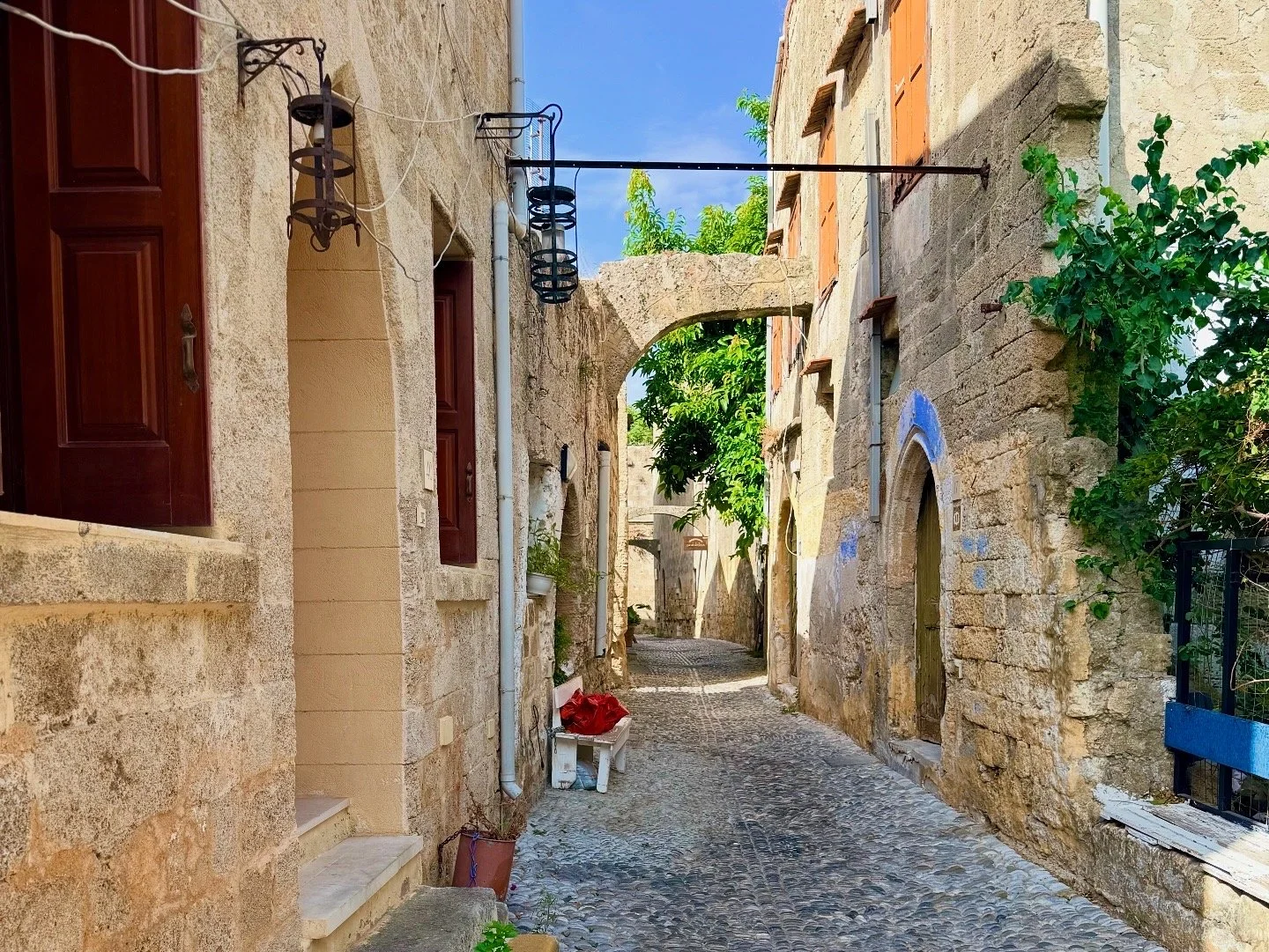 A narrow cobblestone alleyway in a Mediterranean village, with stone buildings on either side, green leafy trees, and a blue sky overhead.