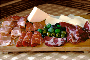 A wooden board with assorted cheeses, slices of prosciutto, salami, fresh blueberries, and a jar of jam.