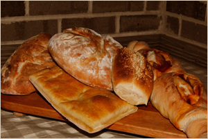 An assortment of bread and baked goods on a wooden tray, including baguettes, rolls, and flatbread, placed against a brick wall background.