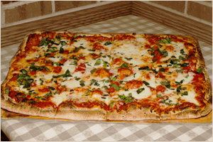 Large rectangular pizza with cheese, tomato sauce, green herbs, and vegetables on a wooden table.