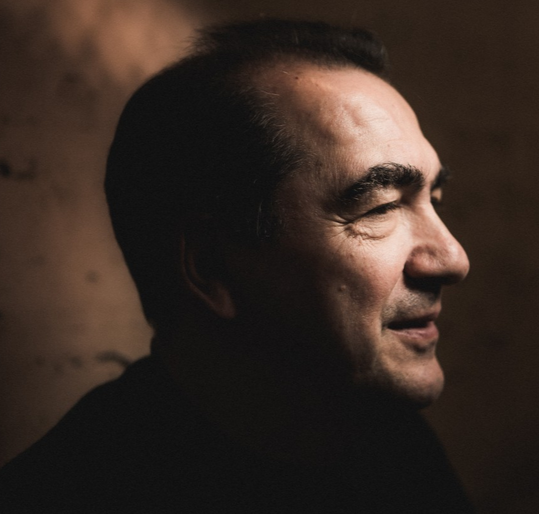 Profile of a smiling middle-aged man with dark hair, viewed from the side, in a dimly lit setting.