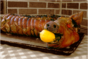 A pig-shaped roasted meat with a yellow lemon in its mouth, displayed on a tray against a brick wall.