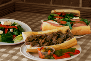 A plate with a beef sandwich, a bowl of salad, and two additional sandwiches with greens and tomatoes in the background.