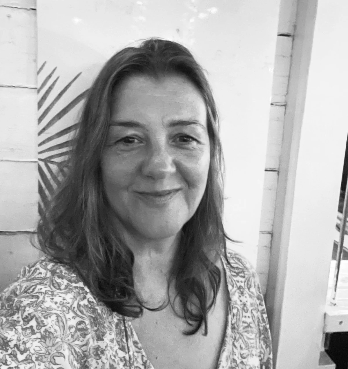 A woman with long, dark hair and earrings stands outdoors in front of a leafy bush, smiling at the camera.
