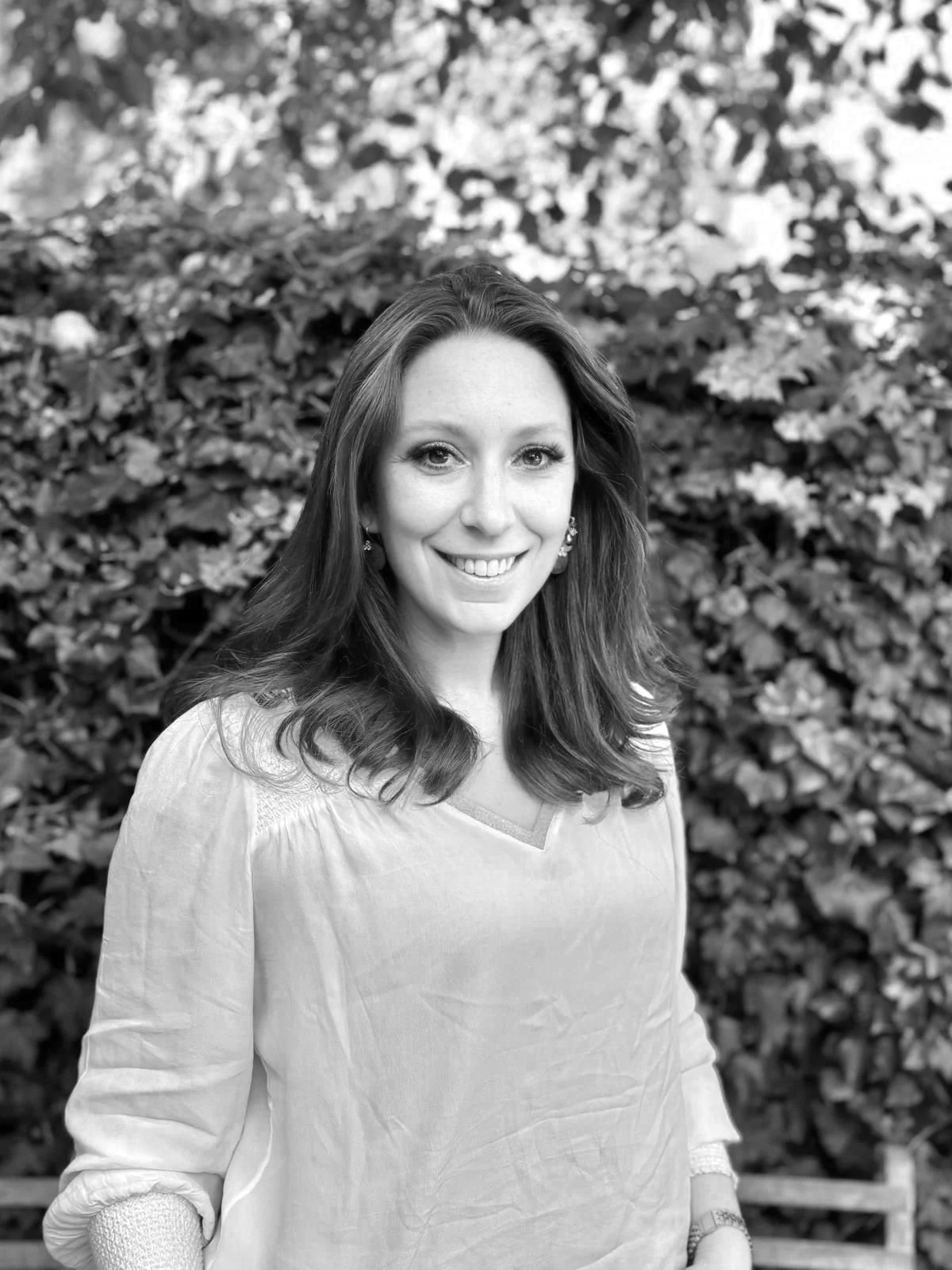 A woman with long hair and a bright smile, standing outdoors in front of a leafy hedge, wearing a light-colored top and earrings.