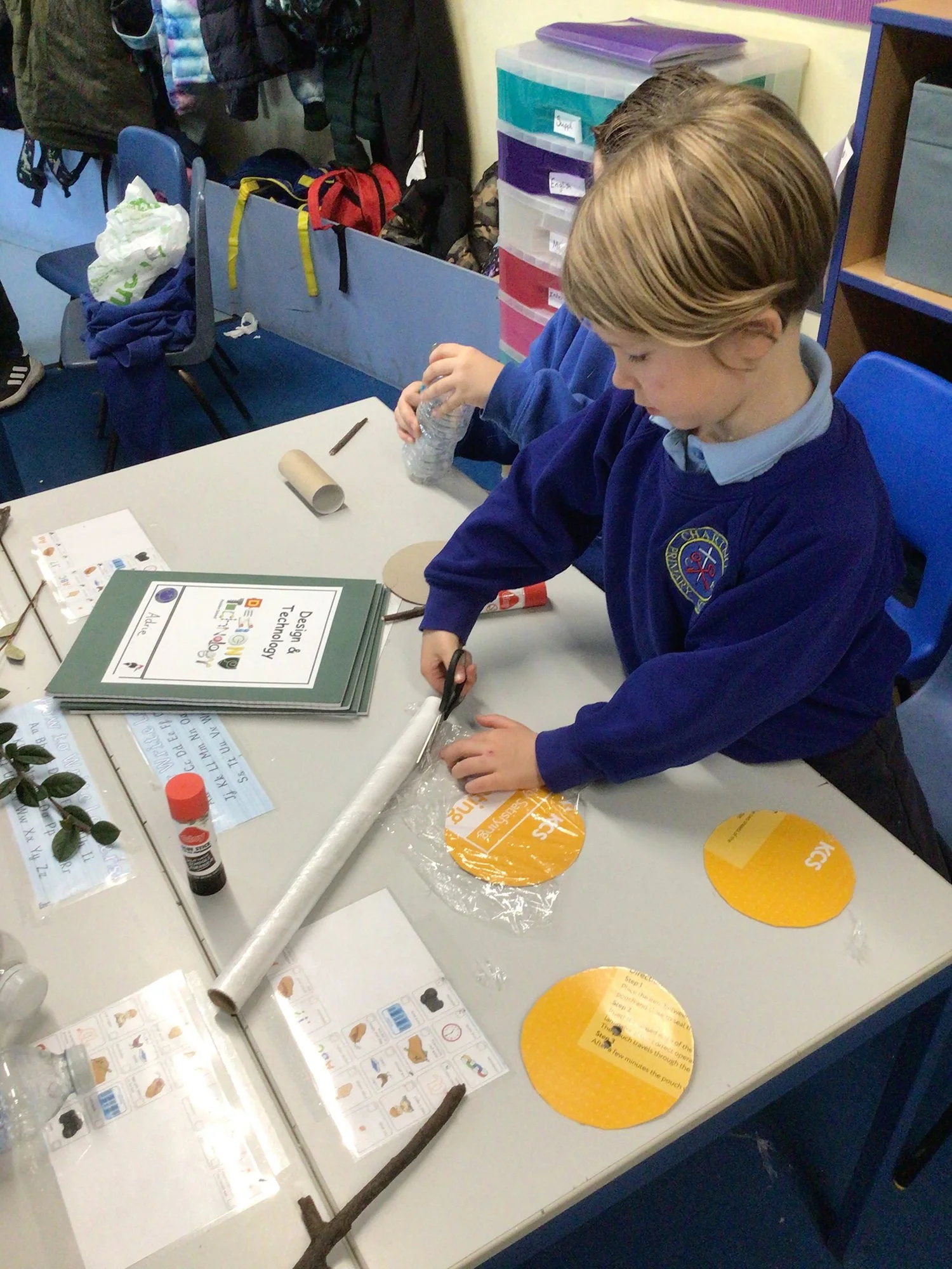 Child in a blue school uniform working on a craft project at a classroom table, using scissors and various craft materials. Books, paper, and supplies are on the table.